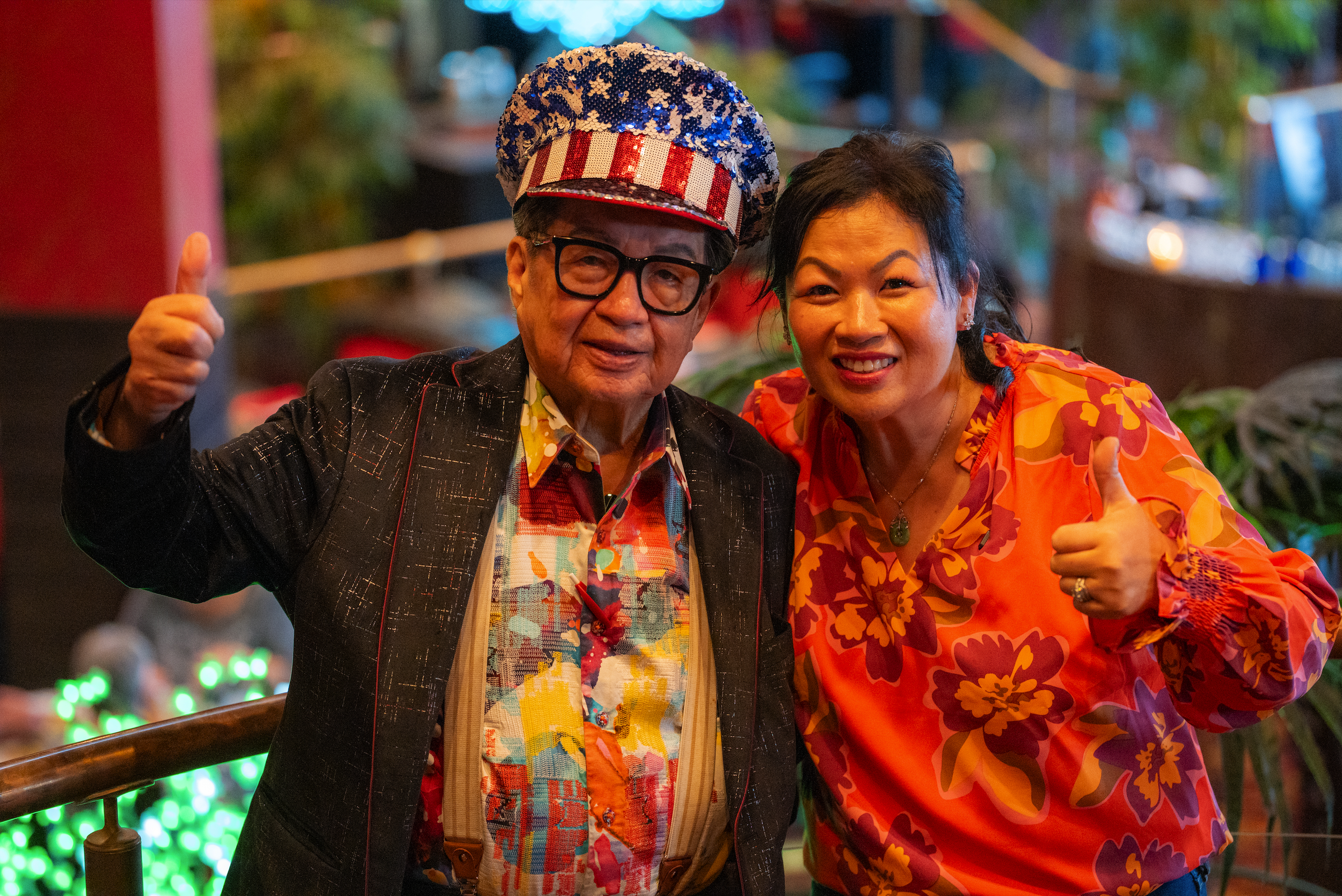 Two people celebrating, giving a thumbs up, wearing colorful and festive outfits with a nighttime, decorated background.
