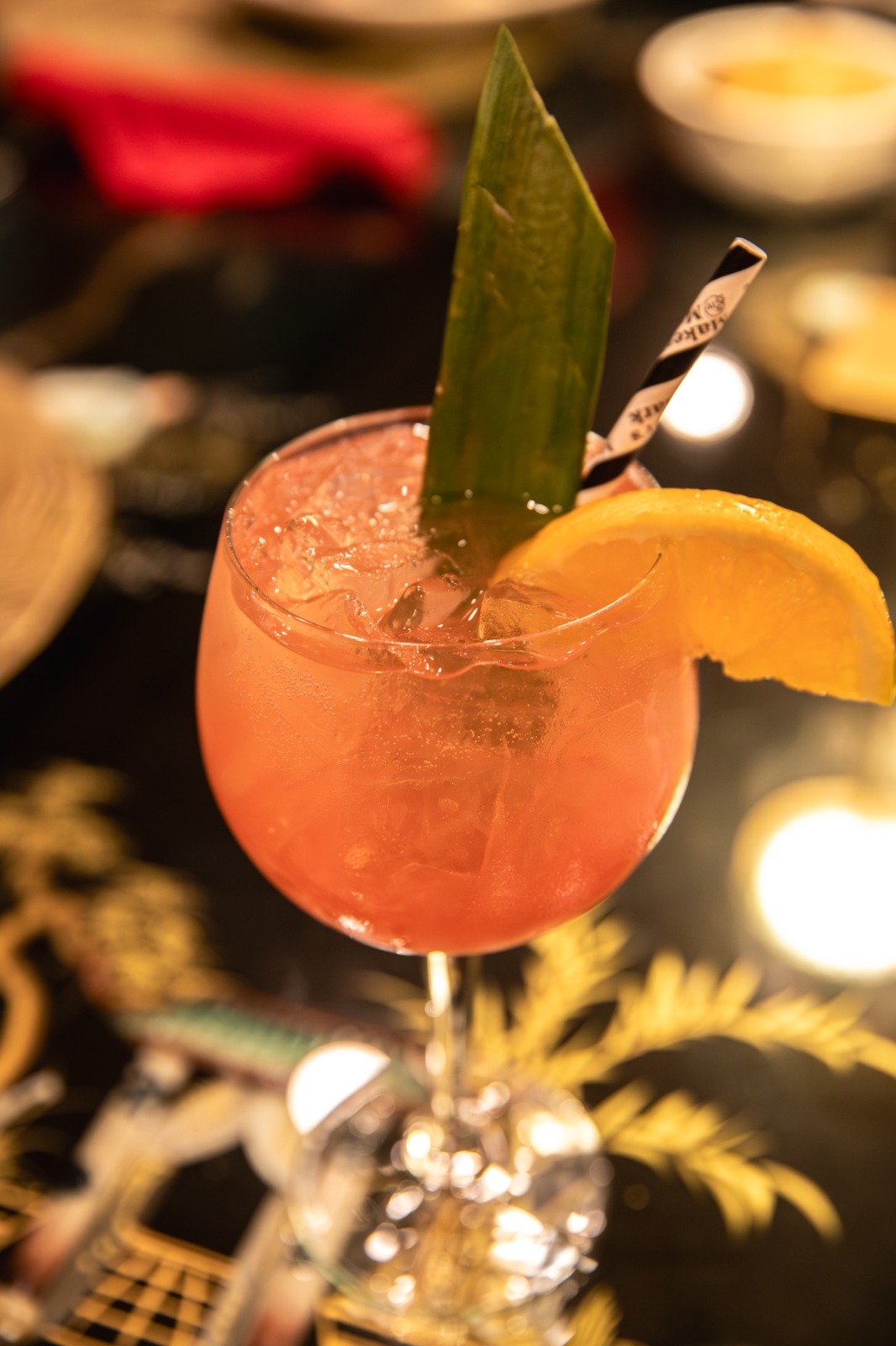 A pink cocktail garnished with a pineapple leaf, an orange slice, and a striped straw, served in a stemmed glass filled with ice.