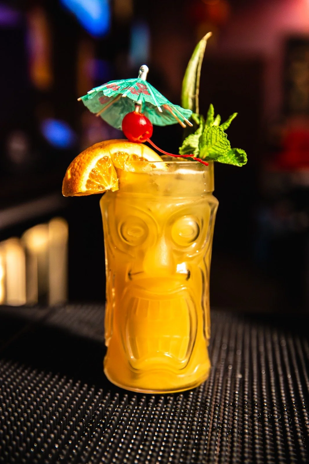 Tiki-themed cocktail in a decorative glass with a face design, garnished with an orange slice, a maraschino cherry, a mint sprig, a small paper umbrella, and a pineapple leaf, on a black bar mat.