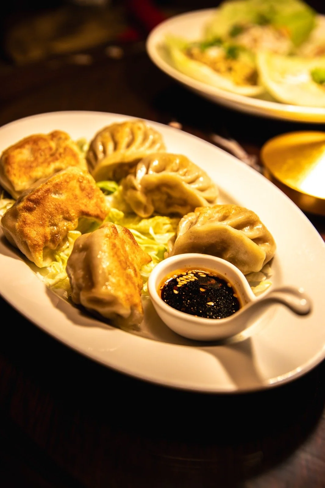 Plate of six fried and steamed dumplings with a small bowl of soy sauce and sesame seeds, garnished with shredded lettuce, on a white oval dish. Carved lemon wedges are partially visible in the background.