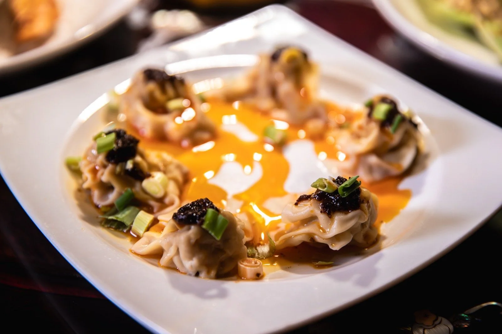A white plate of six lantern-shaped dumplings topped with green onions, black caviar, and a drizzle of hot sauce.