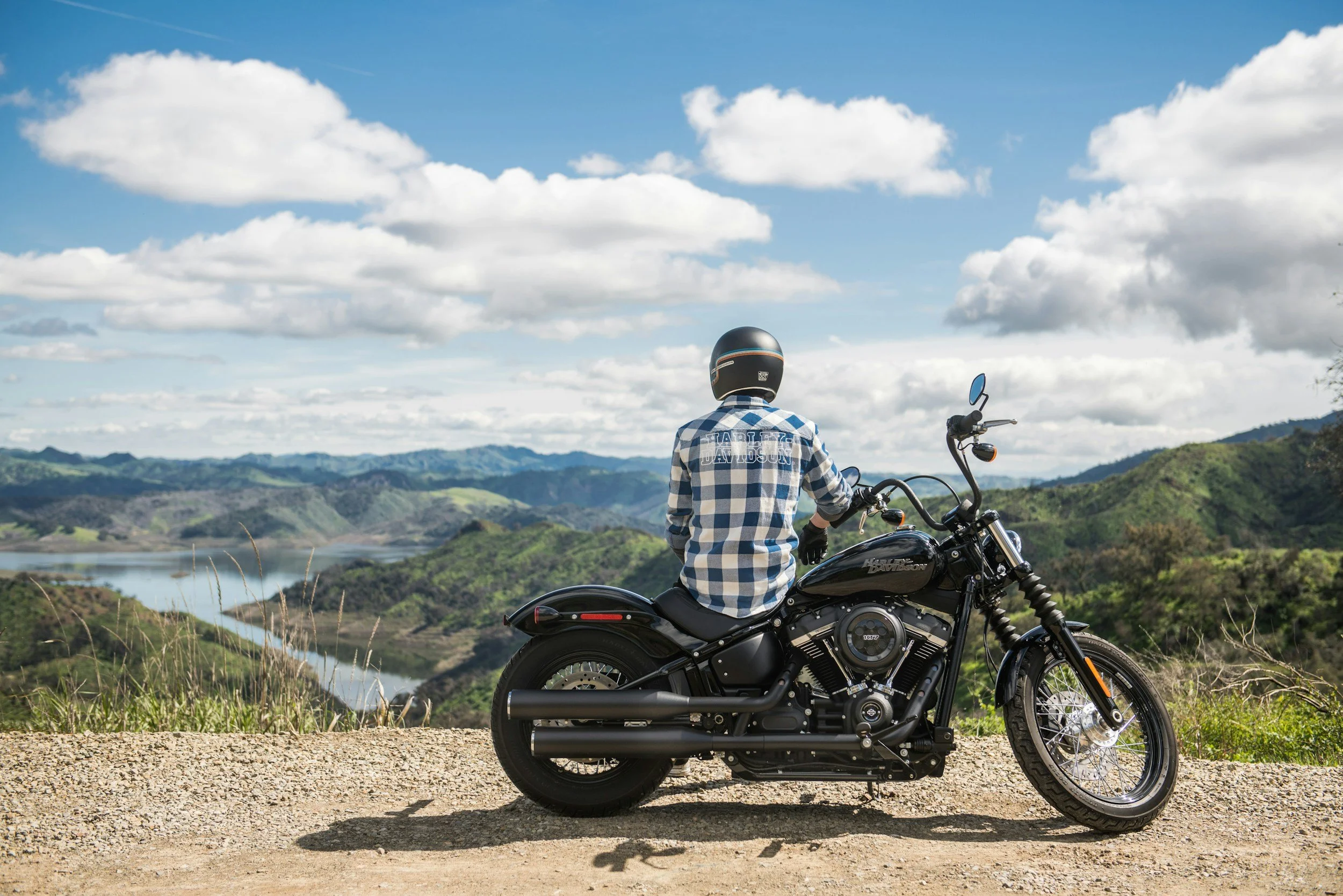 A person wearing a helmet and plaid shirt sitting on a black Harley Davidson motorcycle, overlooking a scenic view of hills, a river, and a sky filled with white clouds.