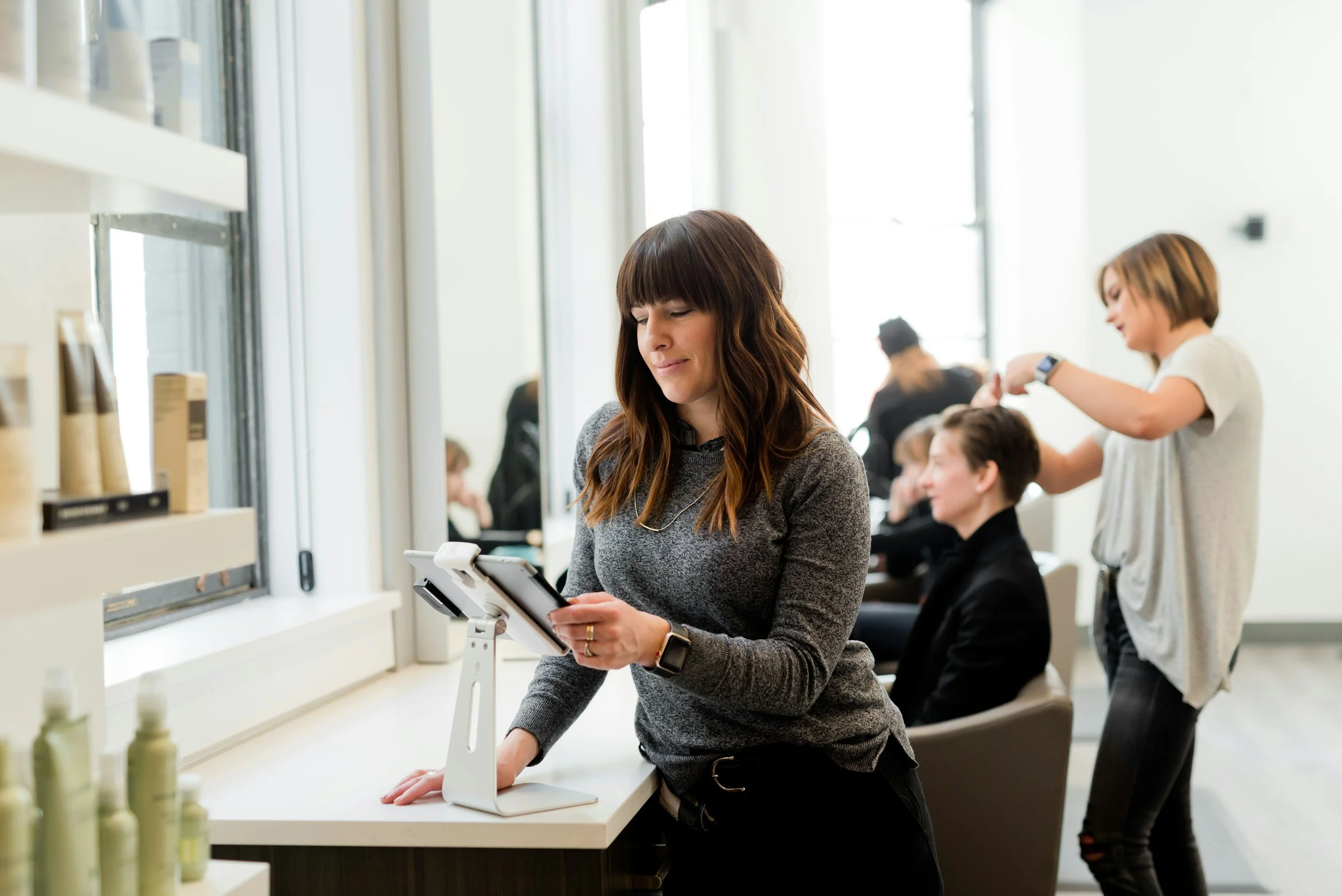 A woman with brown hair and a gray sweater using a digital tablet at a makeup store counter, with a display of skincare bottles nearby. In the background, a woman with short hair in a white shirt is getting her hair styled by a hairstylist, and other customers are seated and standing in a brightly lit modern salon or store.
