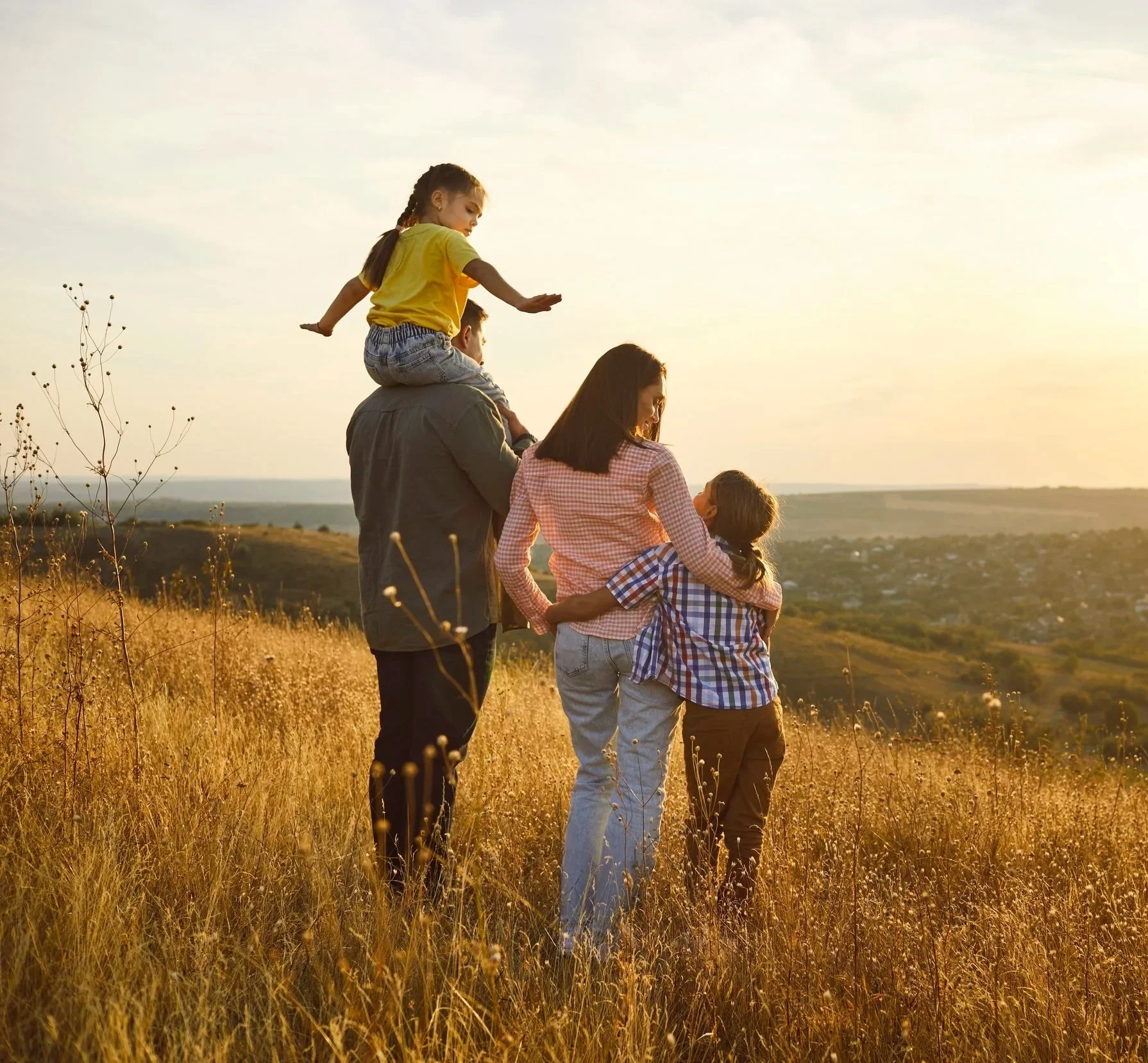 A family walking through a field of tall, golden grass at sunset. The scene includes two adults and three children, with one child sitting on the shoulders of an adult. The sunset sky and distant landscape are visible in the background.
