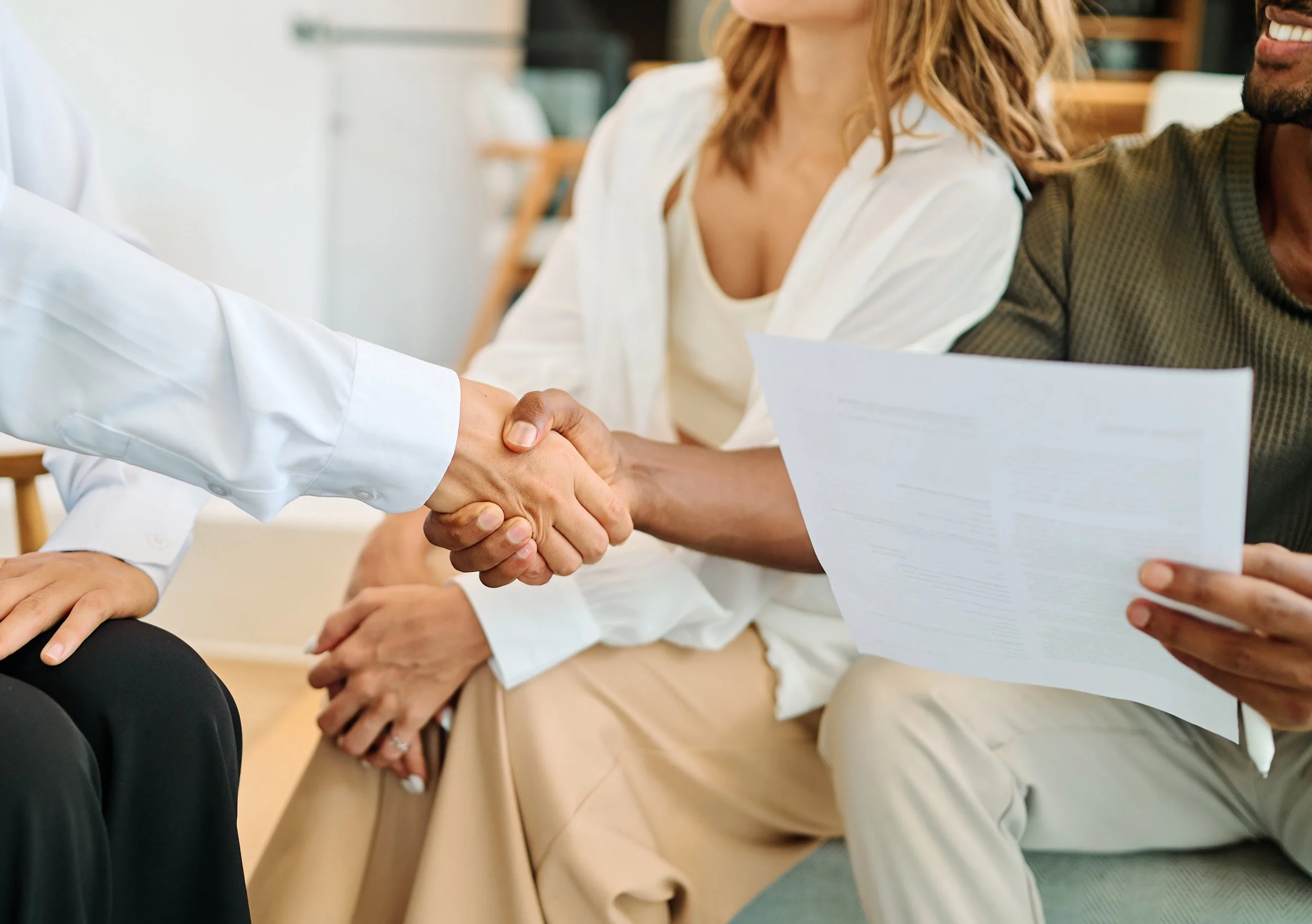 People shaking hands in a professional setting with documents and laptops on a wooden table.