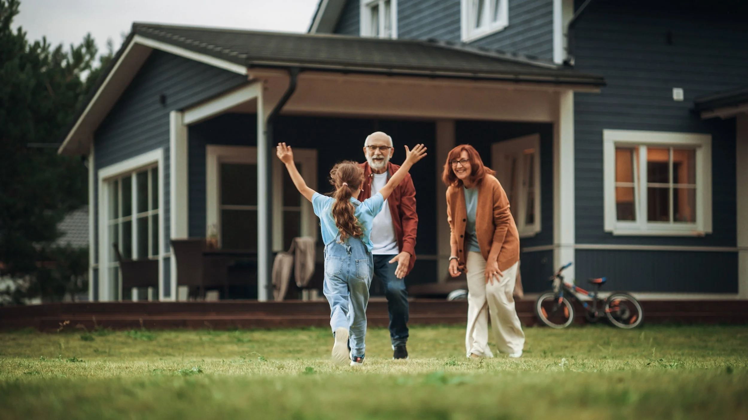 A young girl with long hair, wearing a light blue shirt and overalls, running towards a smiling elderly man and woman in front of a blue house, with a bicycle nearby.