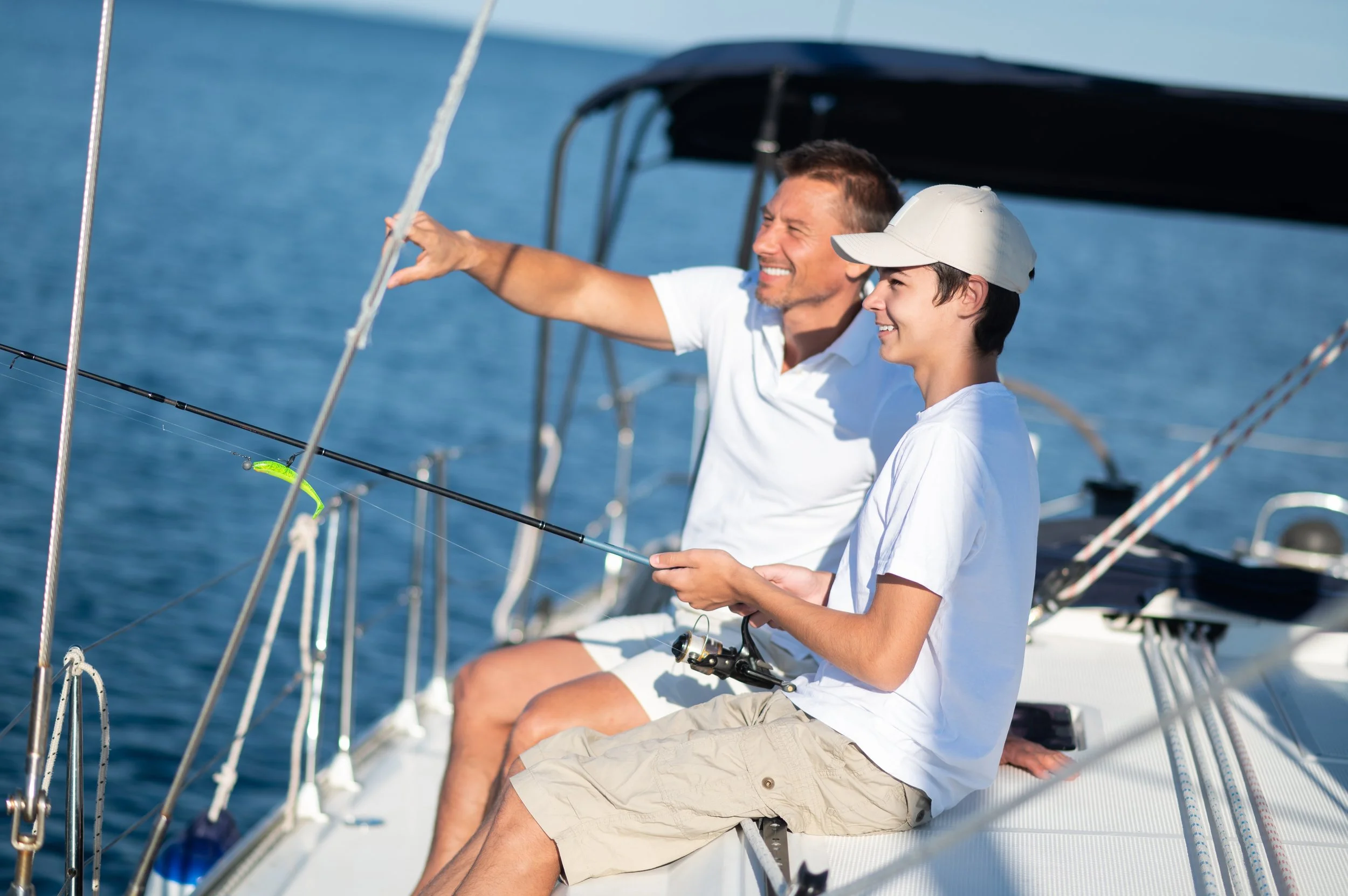Two men sitting on a sailboat, fishing with a rod, enjoying the water and sunny weather.