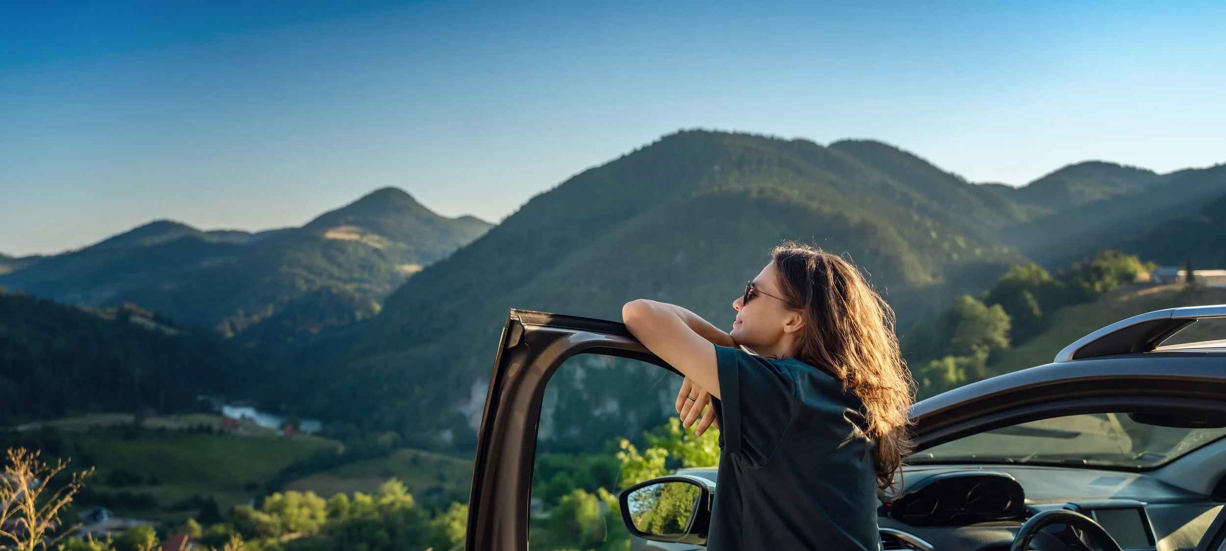 A woman with sunglasses leaning on the open door of a car, looking out at a scenic mountain landscape during sunset.