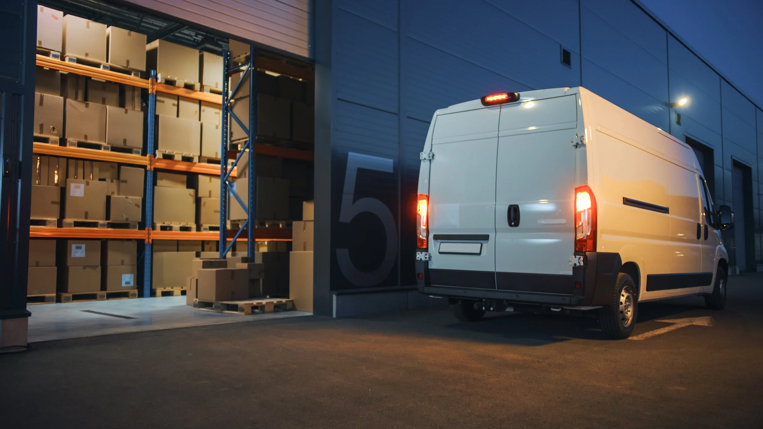 A white delivery van parked outside a warehouse, with shelves of boxes inside that are visible through the open door.