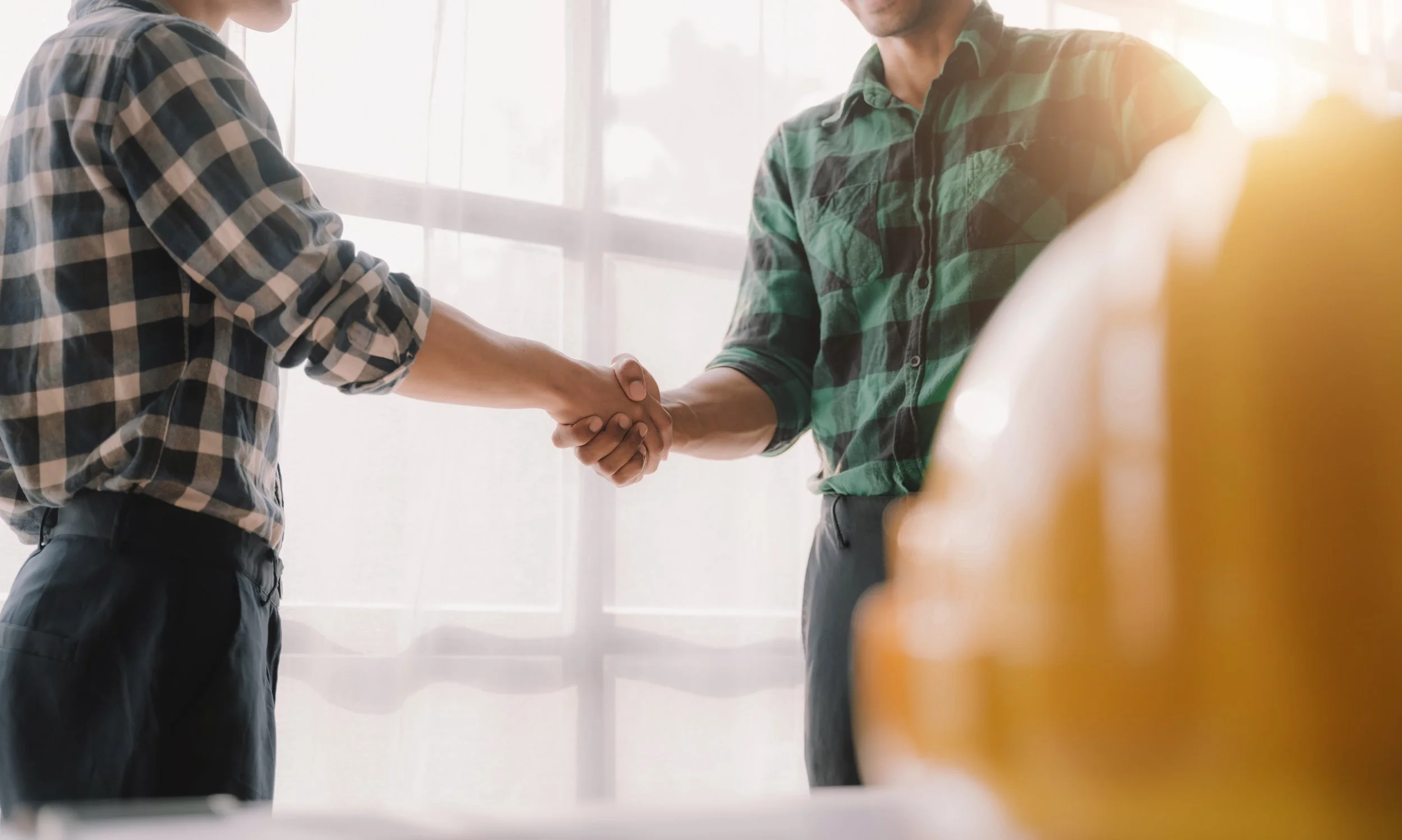 Two men shaking hands indoors near a window, both wearing plaid shirts.
