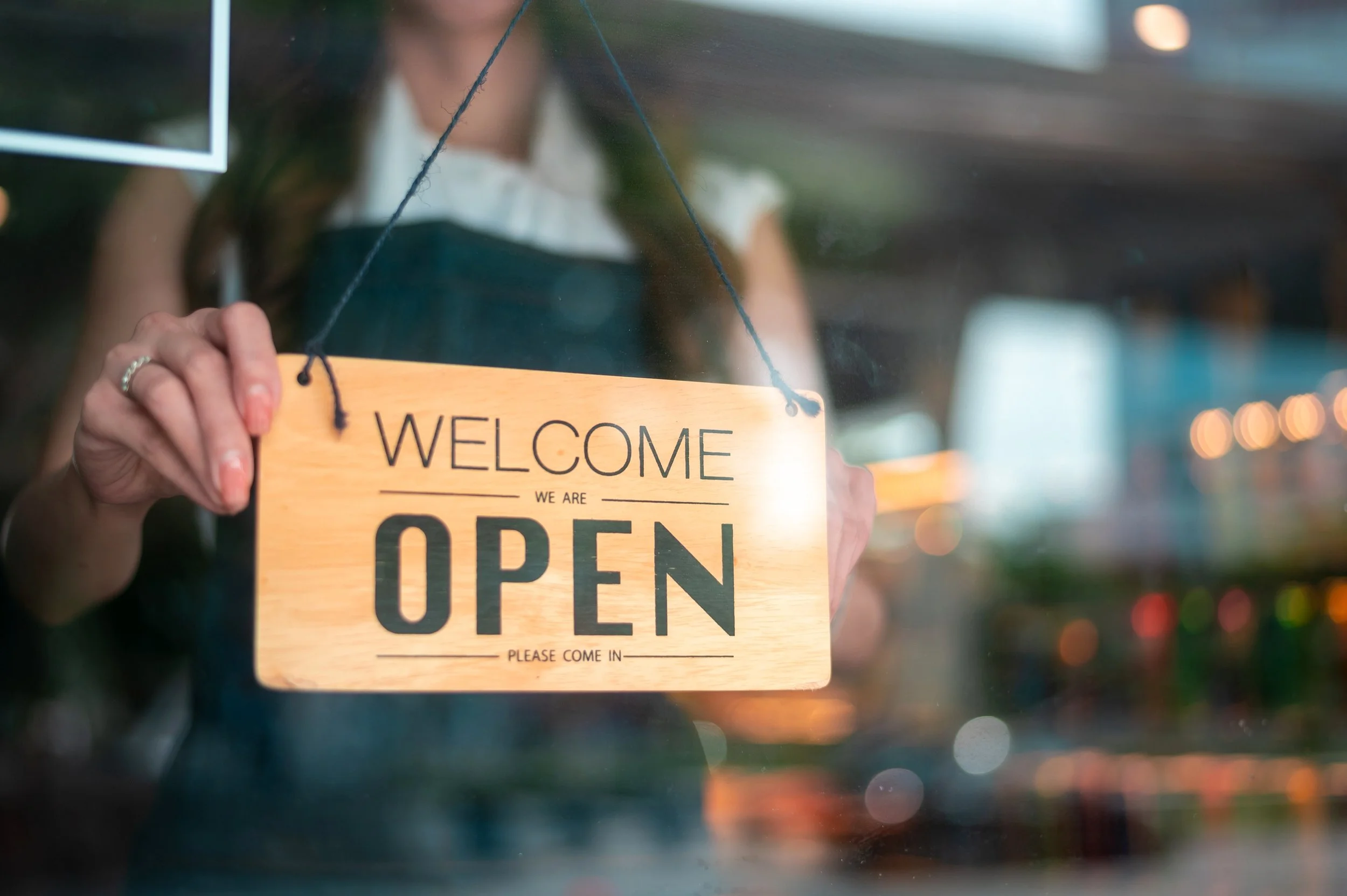 A person is putting up a wooden 'OPEN' sign on a glass door, with the person's hand and a ring visible. The background is blurred with bokeh lights.