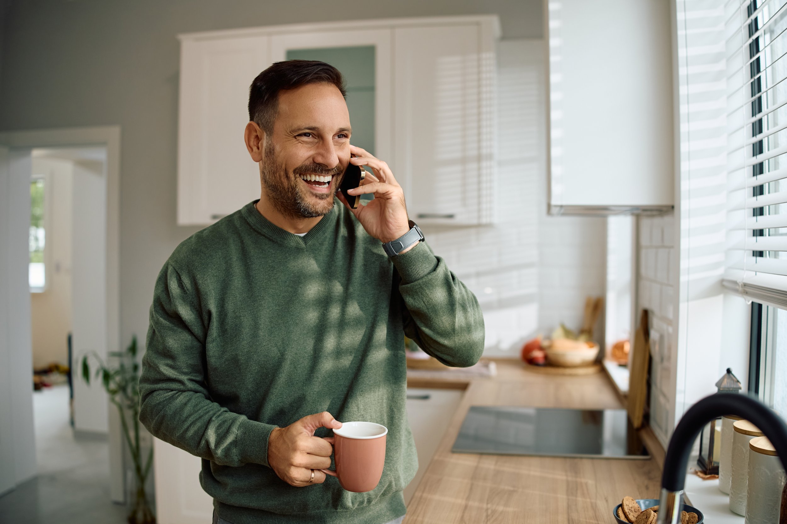 A man smiling and talking on the phone while holding a coffee mug in a modern kitchen with white cabinets and a window with blinds.