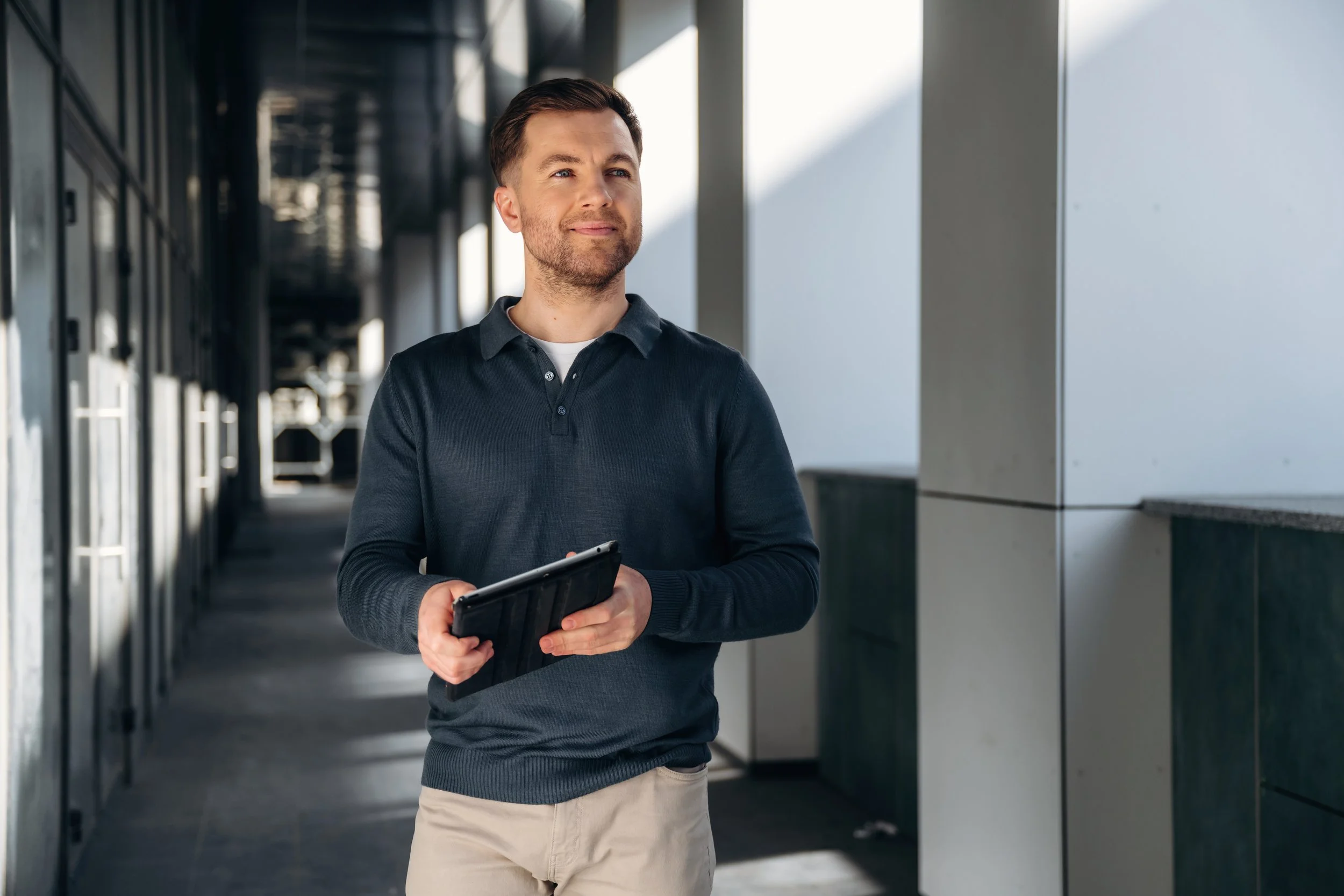 A man holding a tablet in a modern office or corridor with glass walls, bright natural light, and a reflective floor.