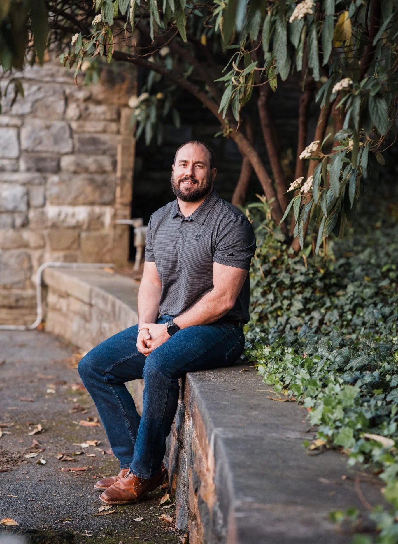 A man with a beard and short hair wearing a short-sleeved gray shirt, blue jeans, and brown shoes sitting on a stone ledge outdoors among green foliage and trees.