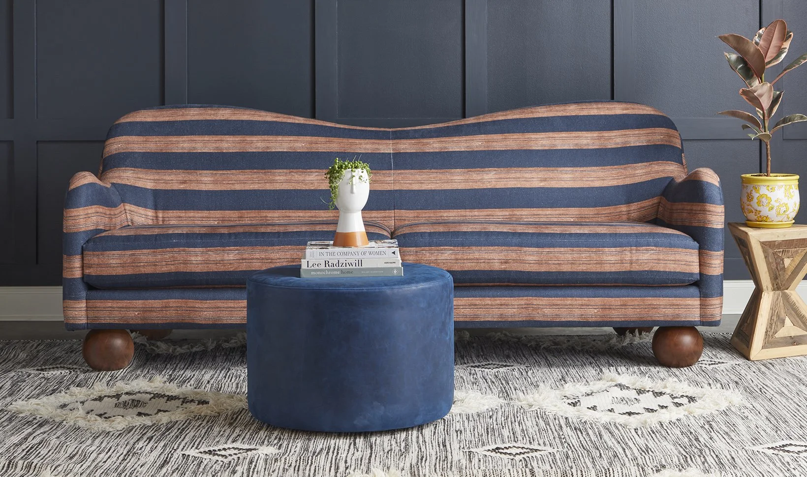 Living room with a striped navy and coral sofa, a blue ottoman with a white vase and books on top, a mustard-colored flowerpot with a plant, and a patterned rug.