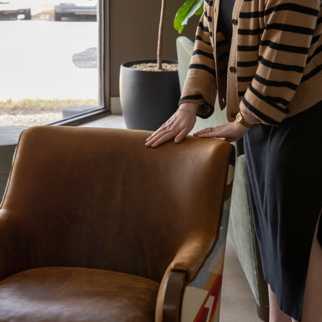 Person leaning on a brown leather armchair, wearing a striped cardigan, near a window with a potted plant nearby.