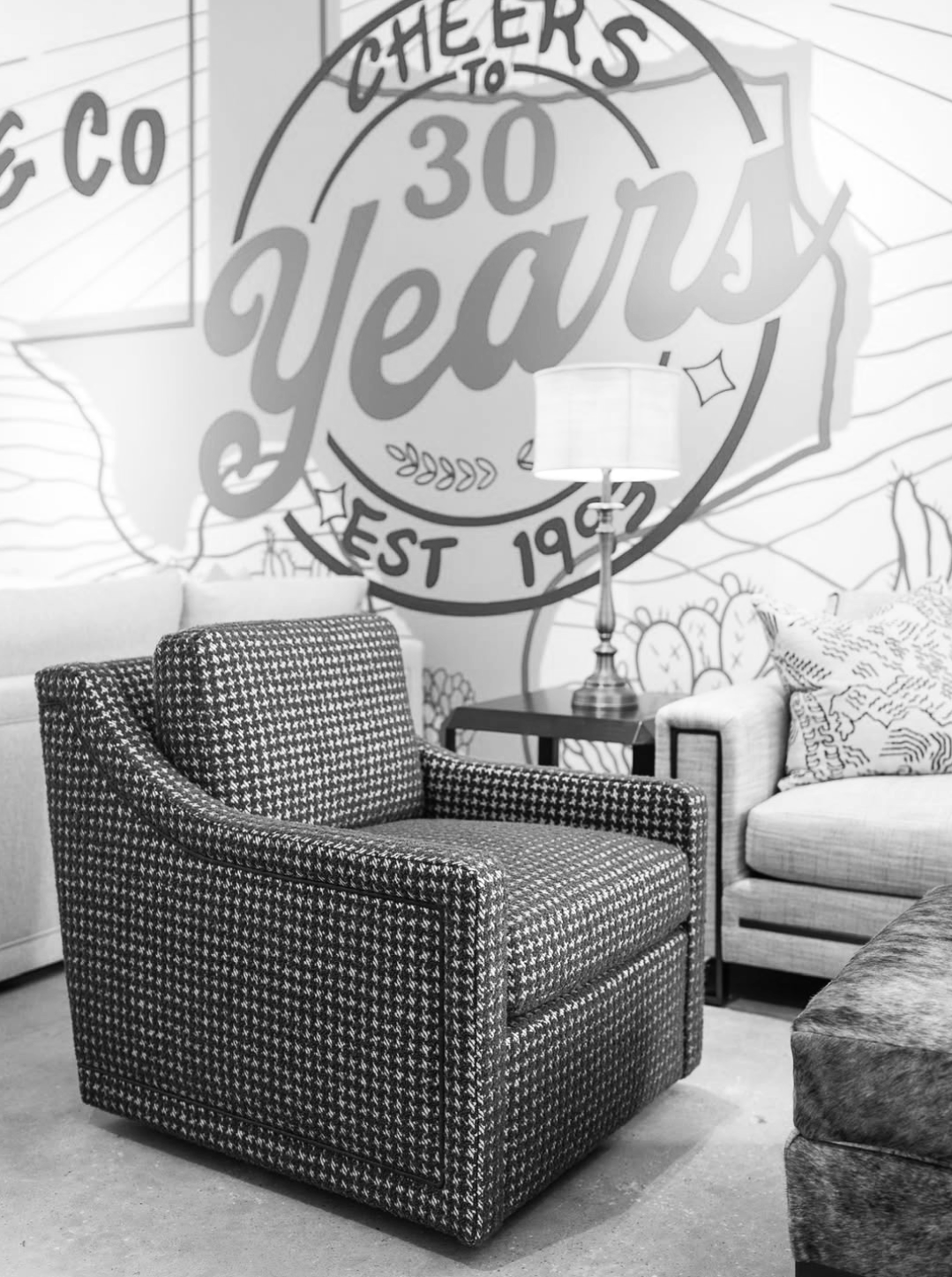 A cozy living room corner with a houndstooth armchair, a side table with a lamp, a patterned pillow on an adjacent sofa, and a mural on the wall celebrating 30 years of cheer and festivities.