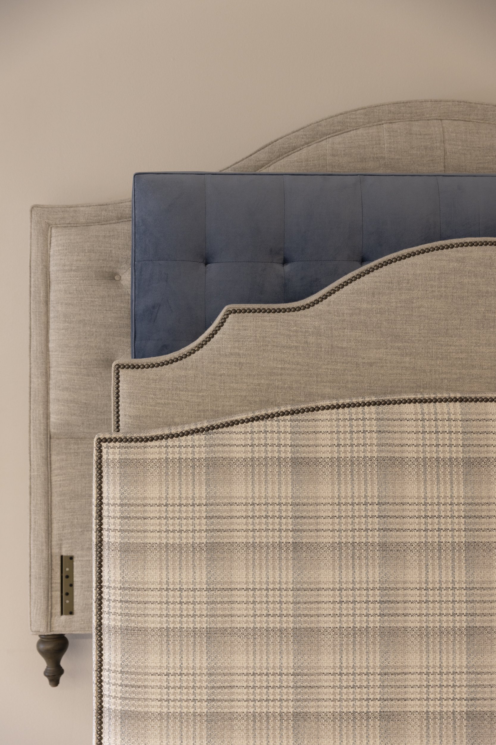 A beige upholstered headboard with nailhead trim, a beige and gray patterned rug, and a partial view of a blue velvet tufted bed or bench.