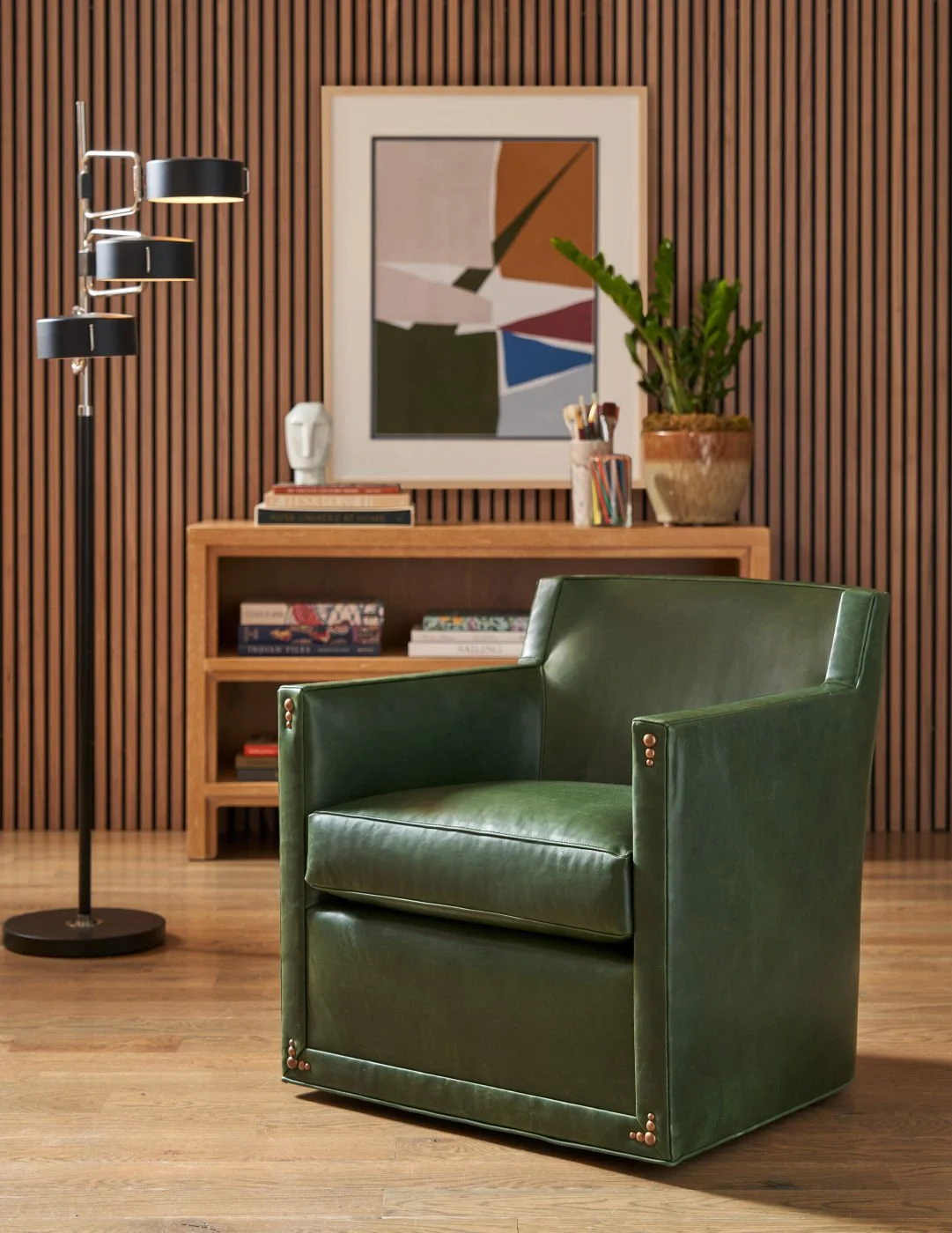 Green leather armchair in a modern room with wooden wall paneling, a wooden bookshelf, framed abstract art, a potted plant, and a floor lamp.