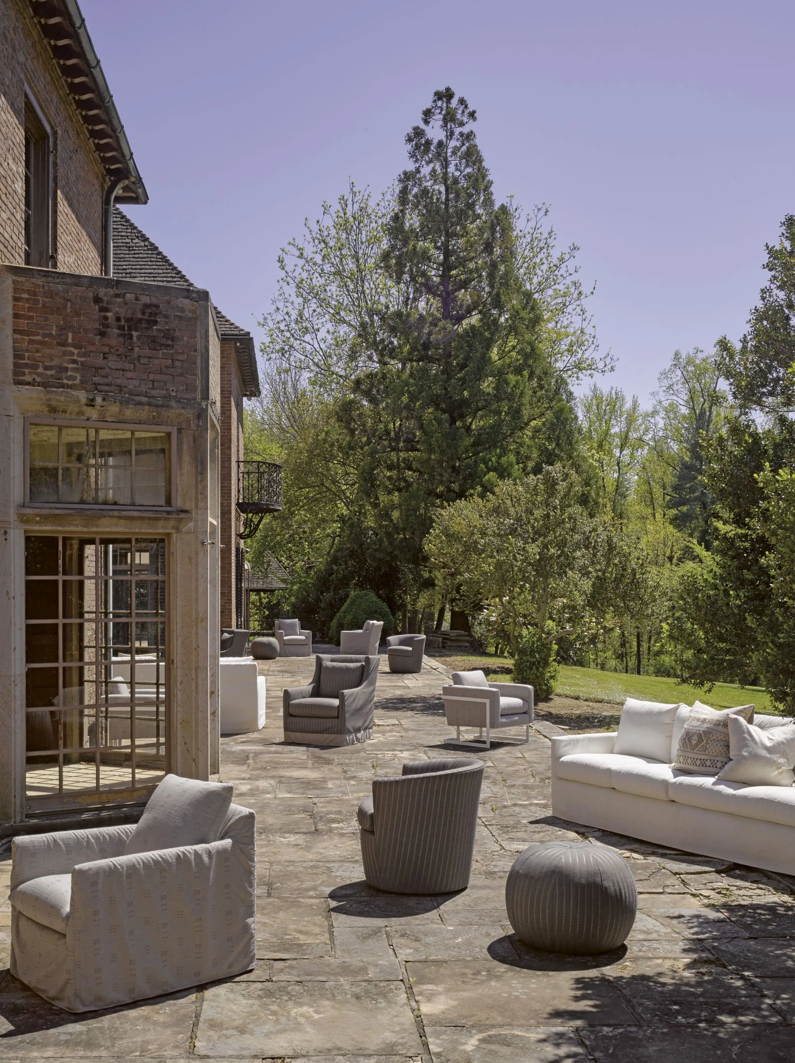 Outdoor patio area with various white and gray chairs and sofas on stone flooring, surrounded by trees and greenery, with a brick house on the left side and a clear blue sky overhead.