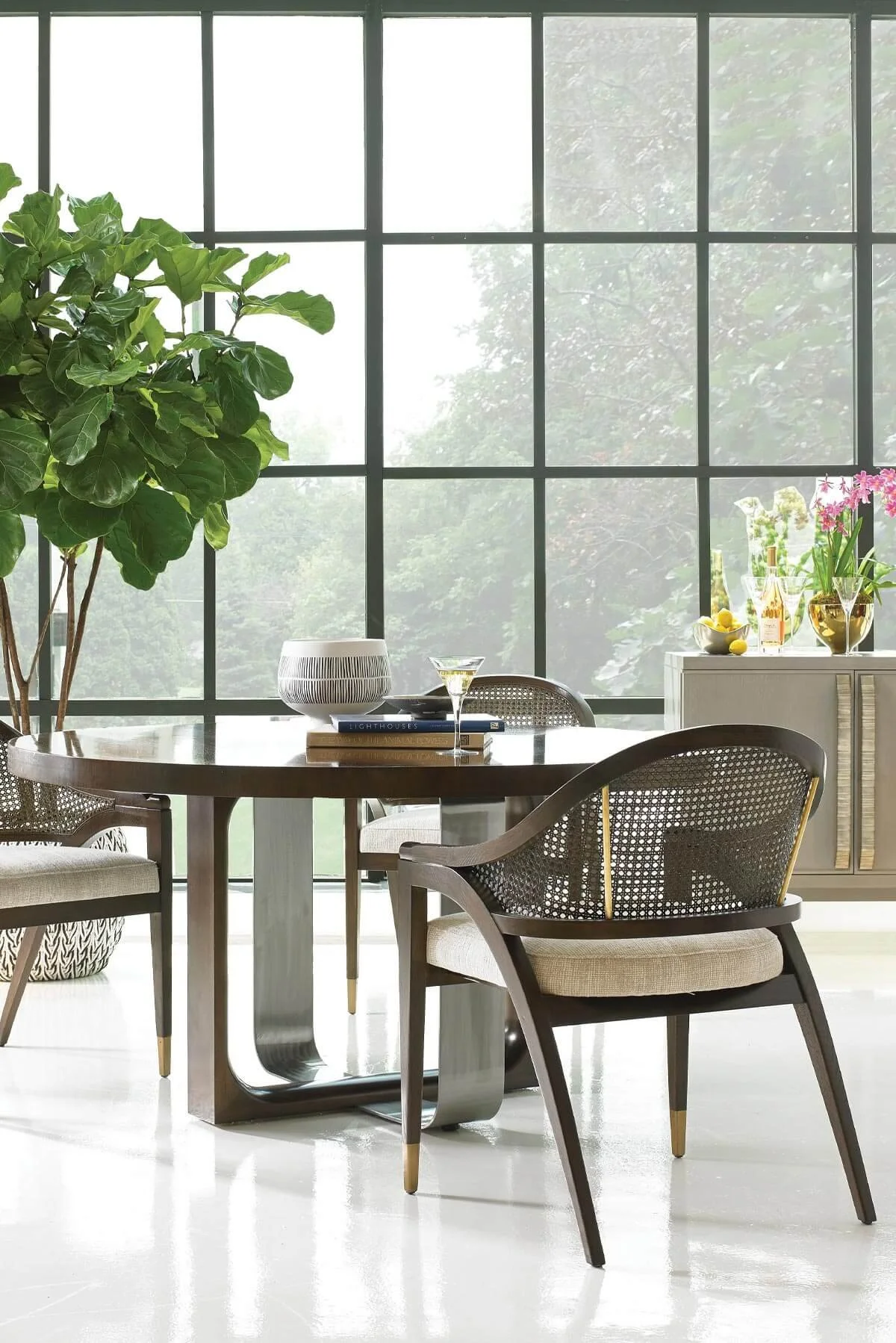 Modern dining area with black round table, chairs with black frames and beige cushions, large window with greenery outside, decorated sideboard with glasses, bottles, fruits, and flowers, large potted plant in corner.