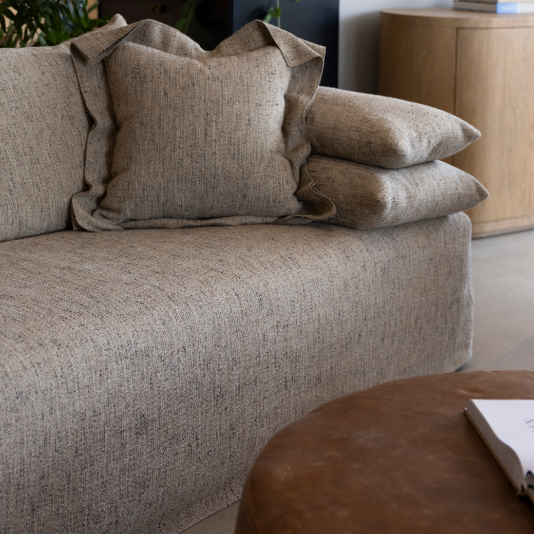 A beige upholstered sofa with a matching cushion and three beige cushions on the right side, next to a wooden sideboard and a round brown coffee table with a book on top.