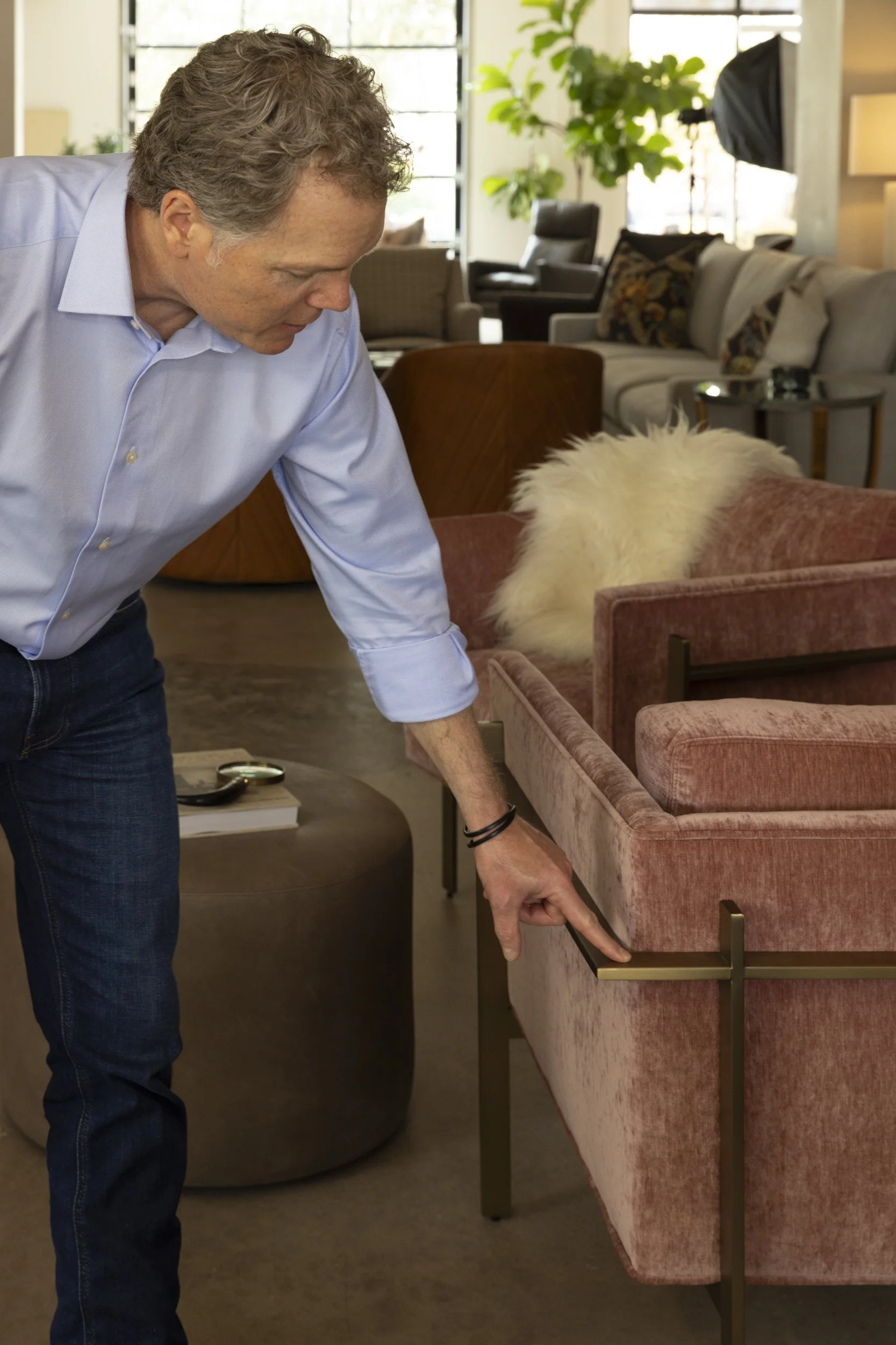 A man in a blue shirt and jeans cleaning or tidying up a pink velvet sofa in a living room with modern furniture and large windows