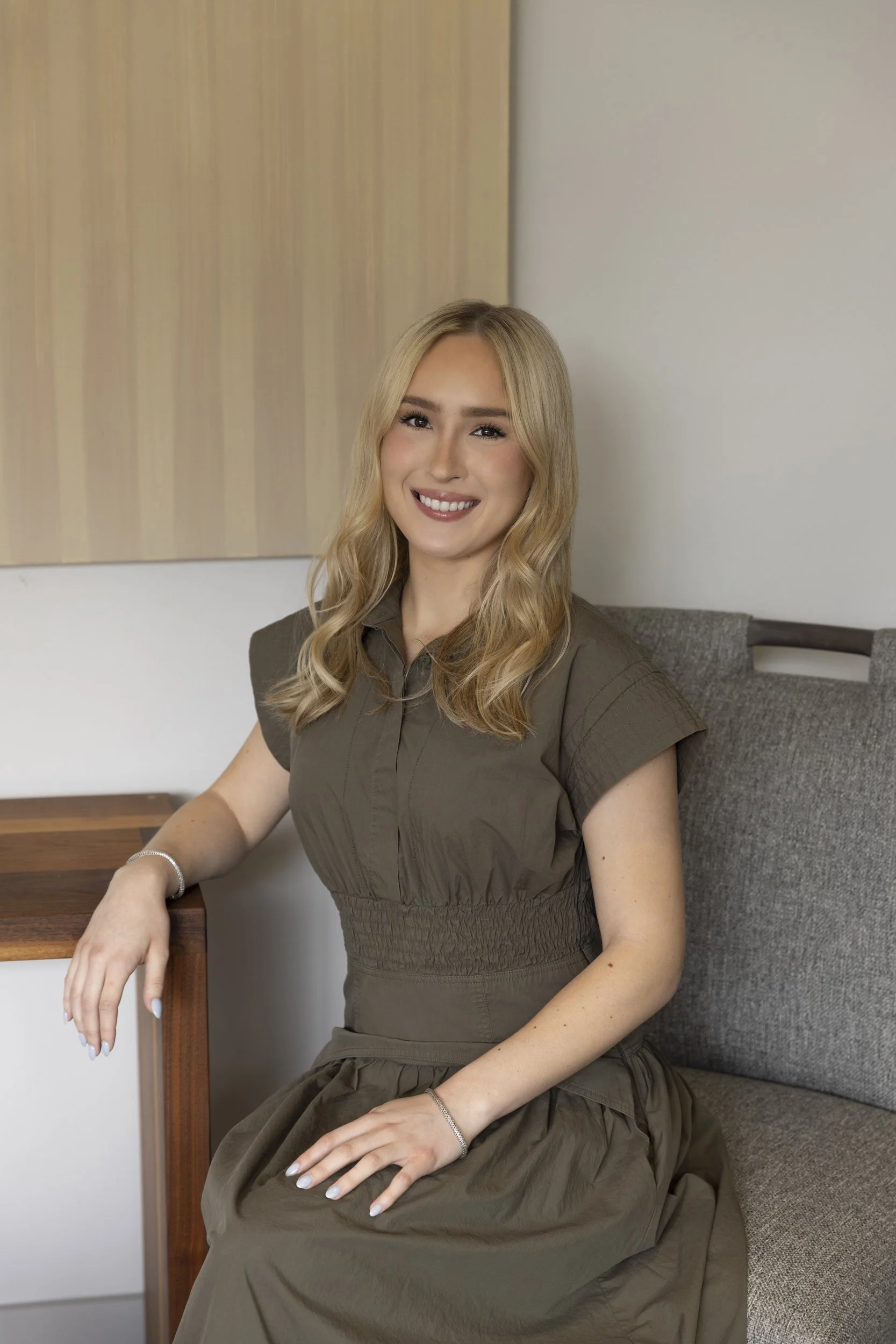 A young woman with long blonde hair, smiling, sitting on a gray couch in a room with beige wall art and a white wall.