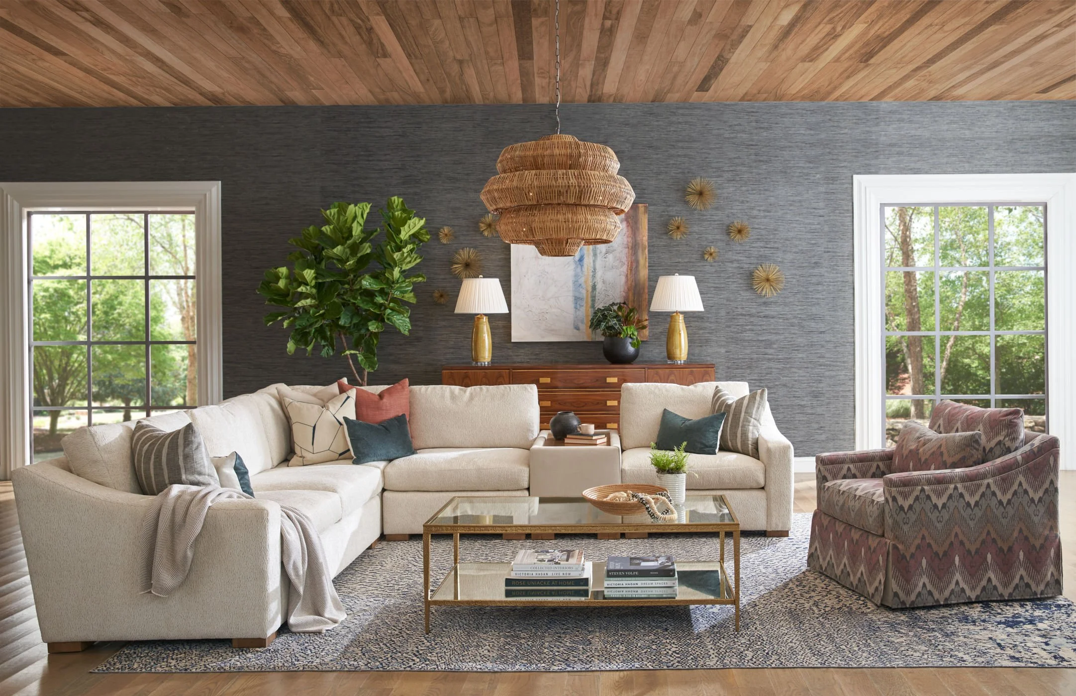 Living room with beige sectional sofa, patterned armchair, glass coffee table, wooden sideboard, large green plant, decorative wall art, wall-mounted sculptures, two large windows with green trees outside, wooden ceiling, and a large wicker pendant light.