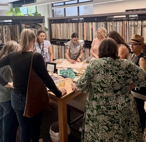 A group of women gathering around a table in a fabric store or showroom, looking at fabric samples and discussing.