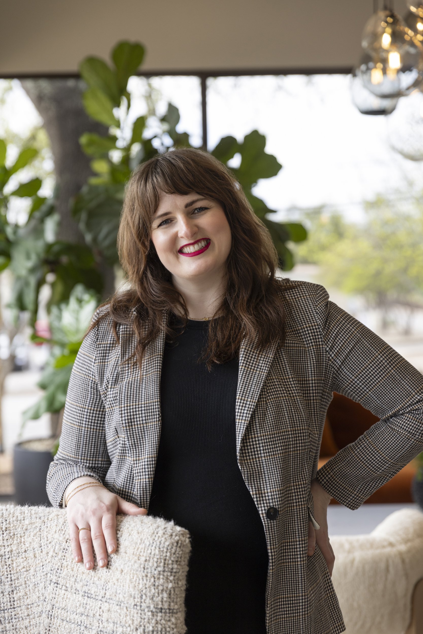 Woman with brown hair and red lipstick smiling, sitting in a cafe with large green plants and a window in the background.