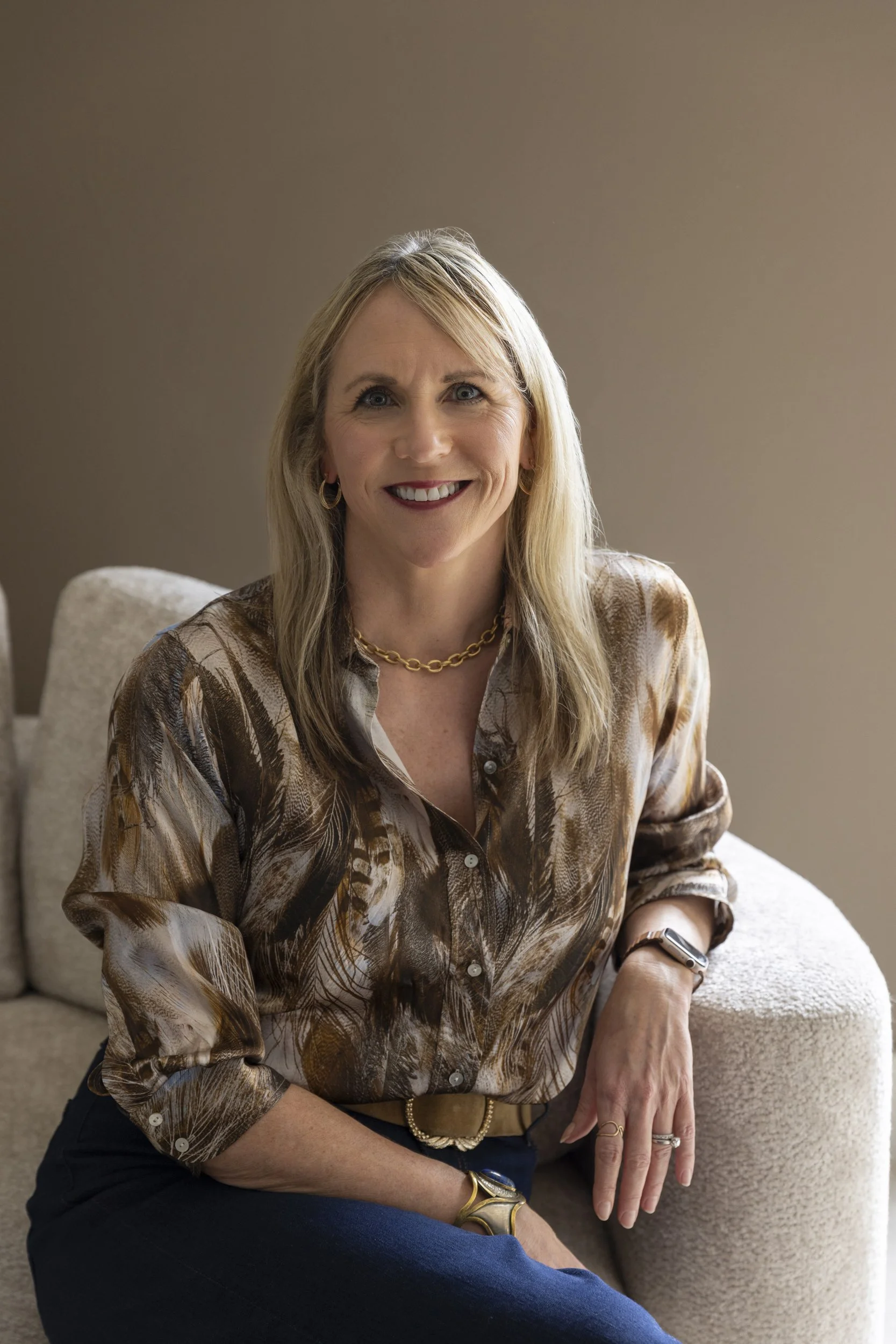 A woman with blonde hair and blue eyes smiling, sitting on a beige couch in a beige room, wearing a patterned blouse, gold jewelry, and watch.
