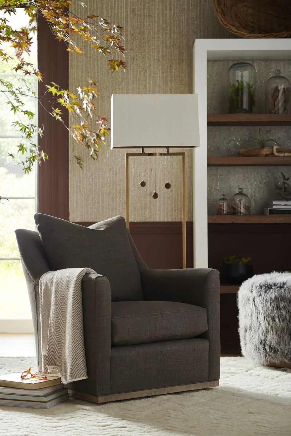Living room with dark grey armchair, beige throw blanket, glass window with greenery outside, beige rug, tall floor lamp with white shade, shelves with decorative items, and a fluffy grey ottoman.