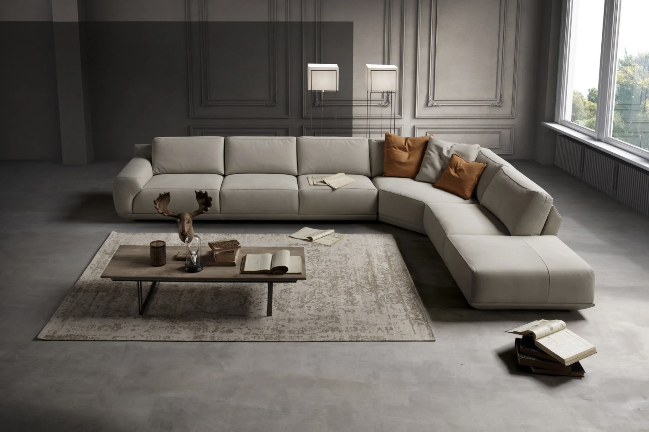 A modern living room with a large L-shaped beige sofa, brown pillows, a wooden coffee table with books, candles, and decorative items, a gray textured wall with molding, tall windows on the right letting in natural light, and a deer head with antlers on the coffee table.