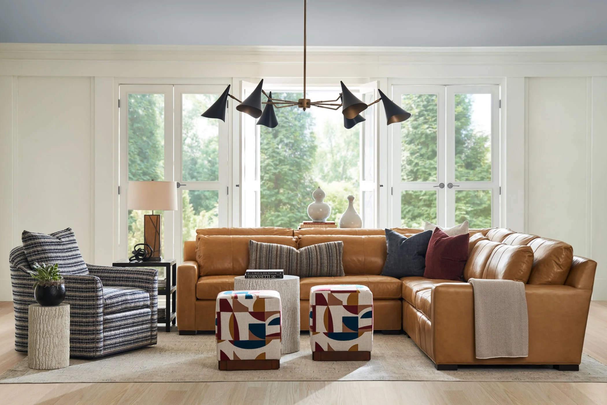 A living room with a tan leather sectional, decorative pillows, a patterned armchair, a black side table with a black vase and green plant, a lamp, and a modern black chandelier hanging from the ceiling. Large windows with a view of green trees in the background.