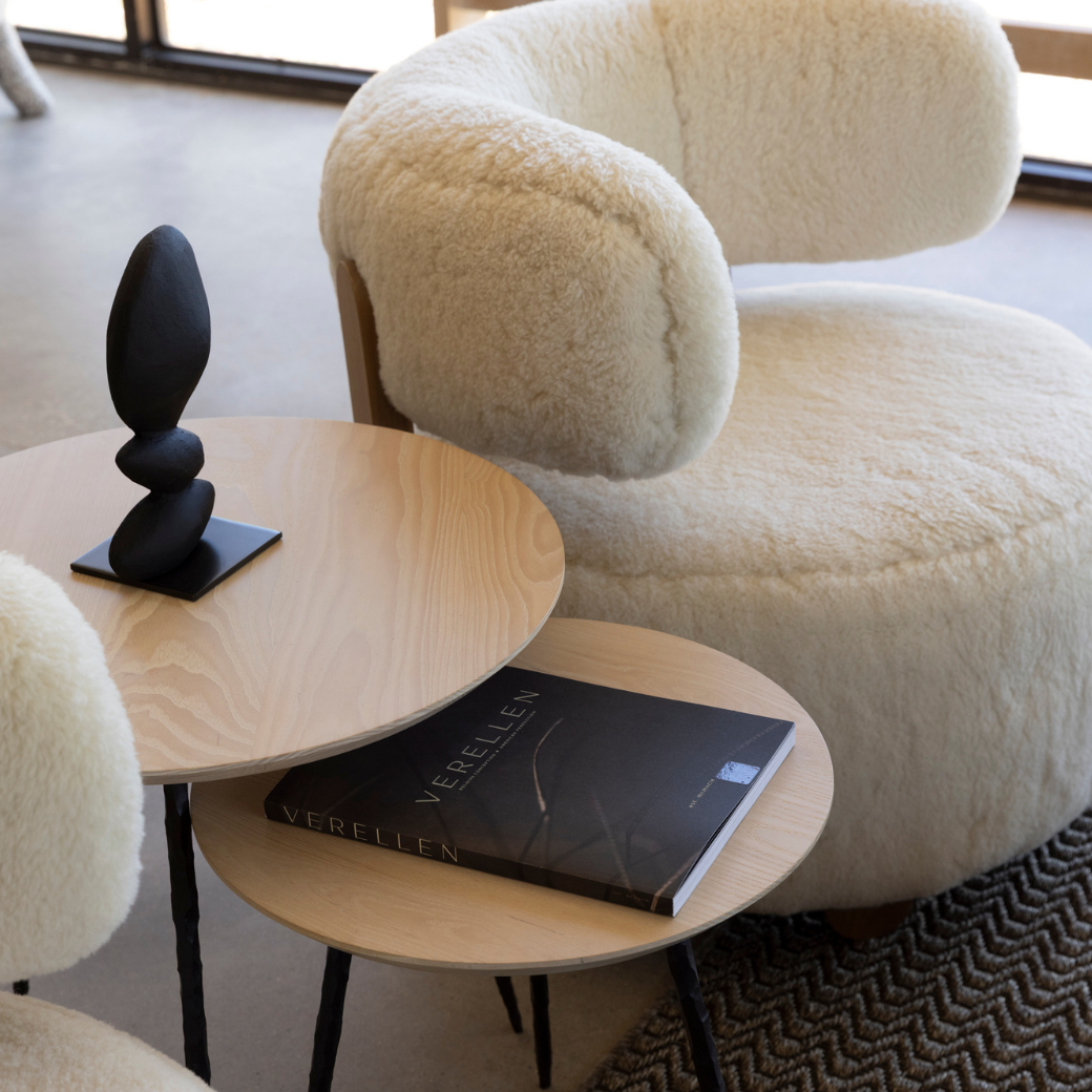 A cozy seating area with a plush, cream-colored armchair, two round wooden side tables stacked together with a black sculpture on the top table and a magazine titled 'Verellen' on the lower table, near a large window letting in natural light.