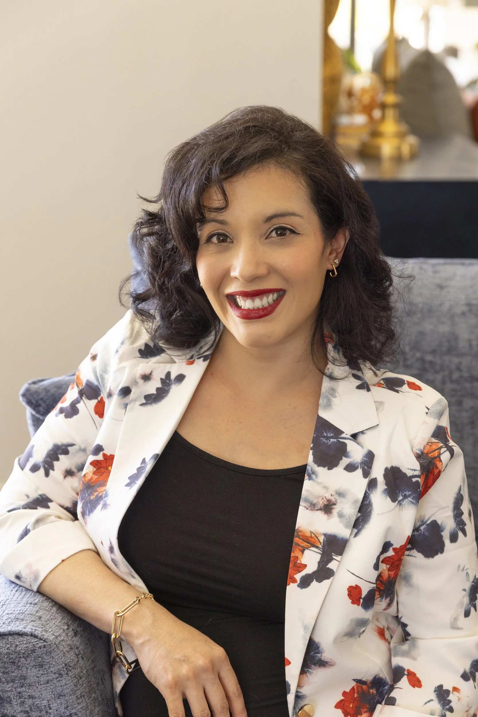 A woman with dark curly hair and red lipstick smiling, dressed in a black shirt and a white floral blazer, sitting on a gray chair with a blurred background of a room with a dark cabinet, lamps, and decorative items.