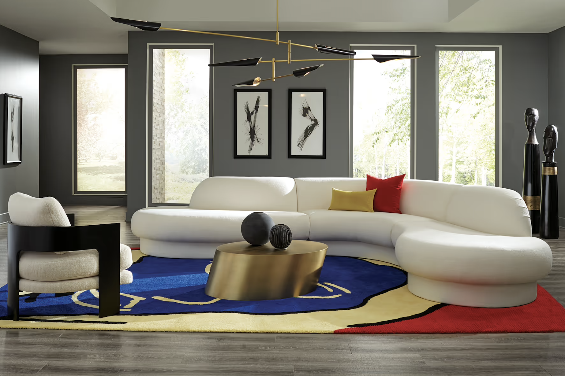 Modern living room with large white curved sectional sofa, a small round gold coffee table with black vases, a blue and multicolor area rug, black and white artwork on dark gray walls, and large windows with greenery outside. The room features a contemporary black and gold chandelier and tall black decorative sculptures.