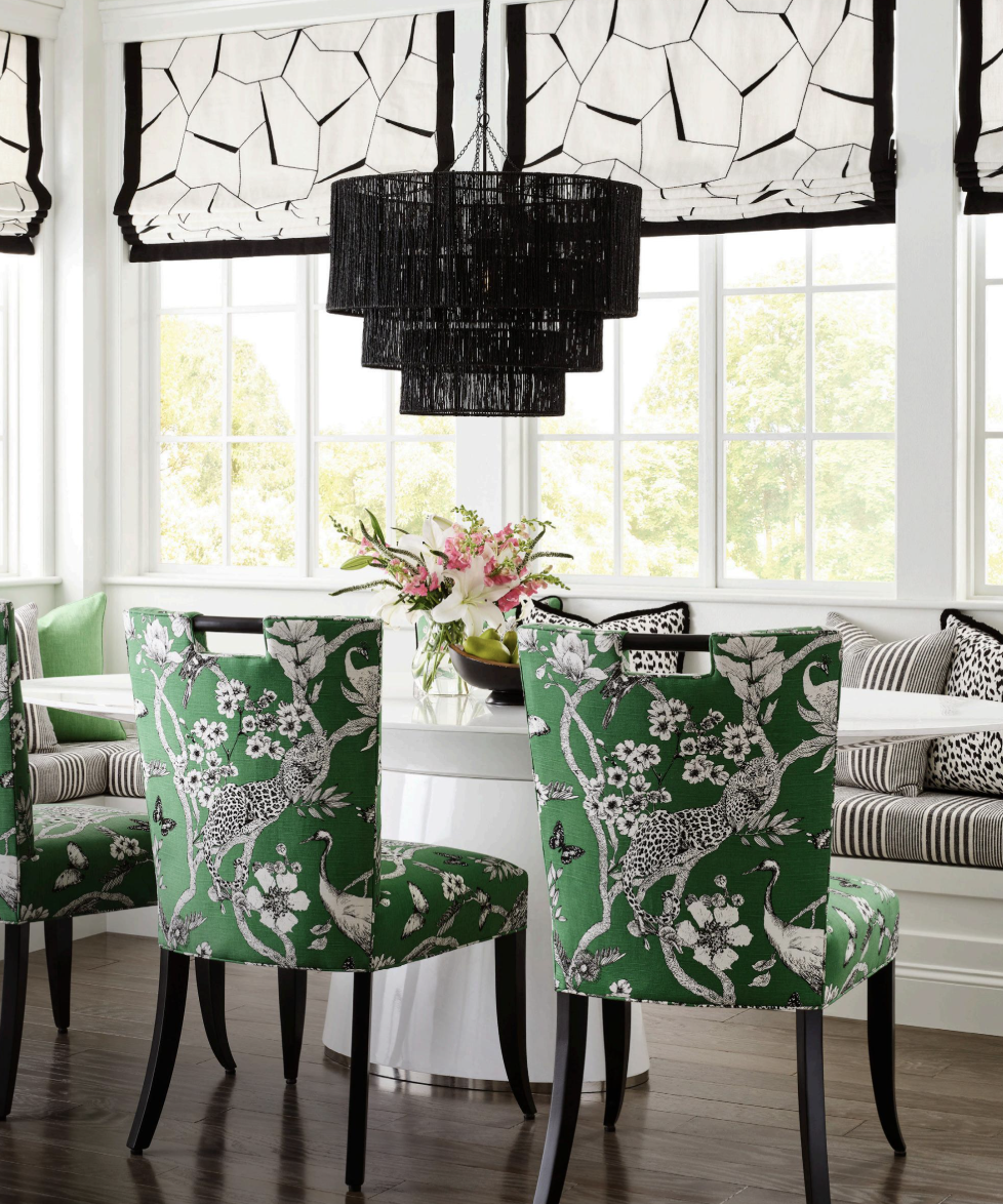 Dining room with green and black patterned chairs, a white round table with a floral centerpiece, large windows with black and white geometric curtains, and a black tiered chandelier.