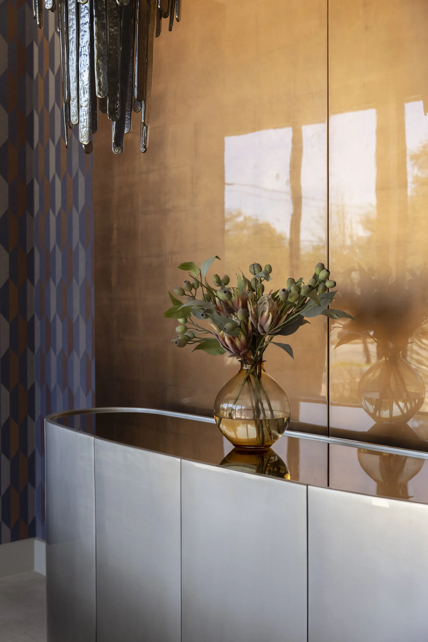 A decorative flower arrangement in a clear glass vase with water, placed on a curved white cabinet in front of a wooden wall with a reflective surface. A mirrored reflection of the vase and some trees are visible on the wall.