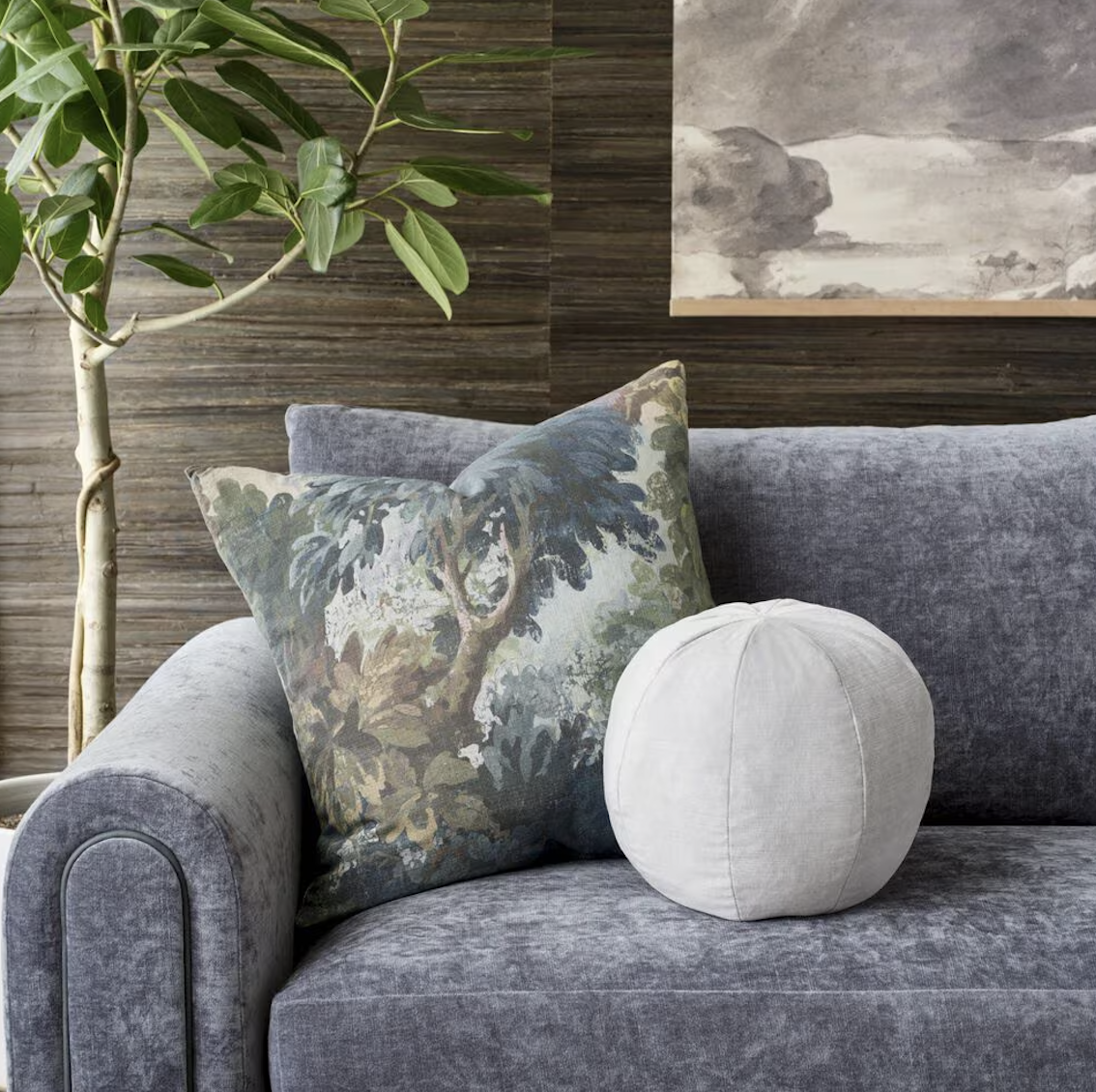 A cozy living room corner with a gray velvet sofa, a decorative pillow with an abstract tree design, a round white cushion, a green leafy plant, and a wooden wall with a framed black and white landscape artwork.