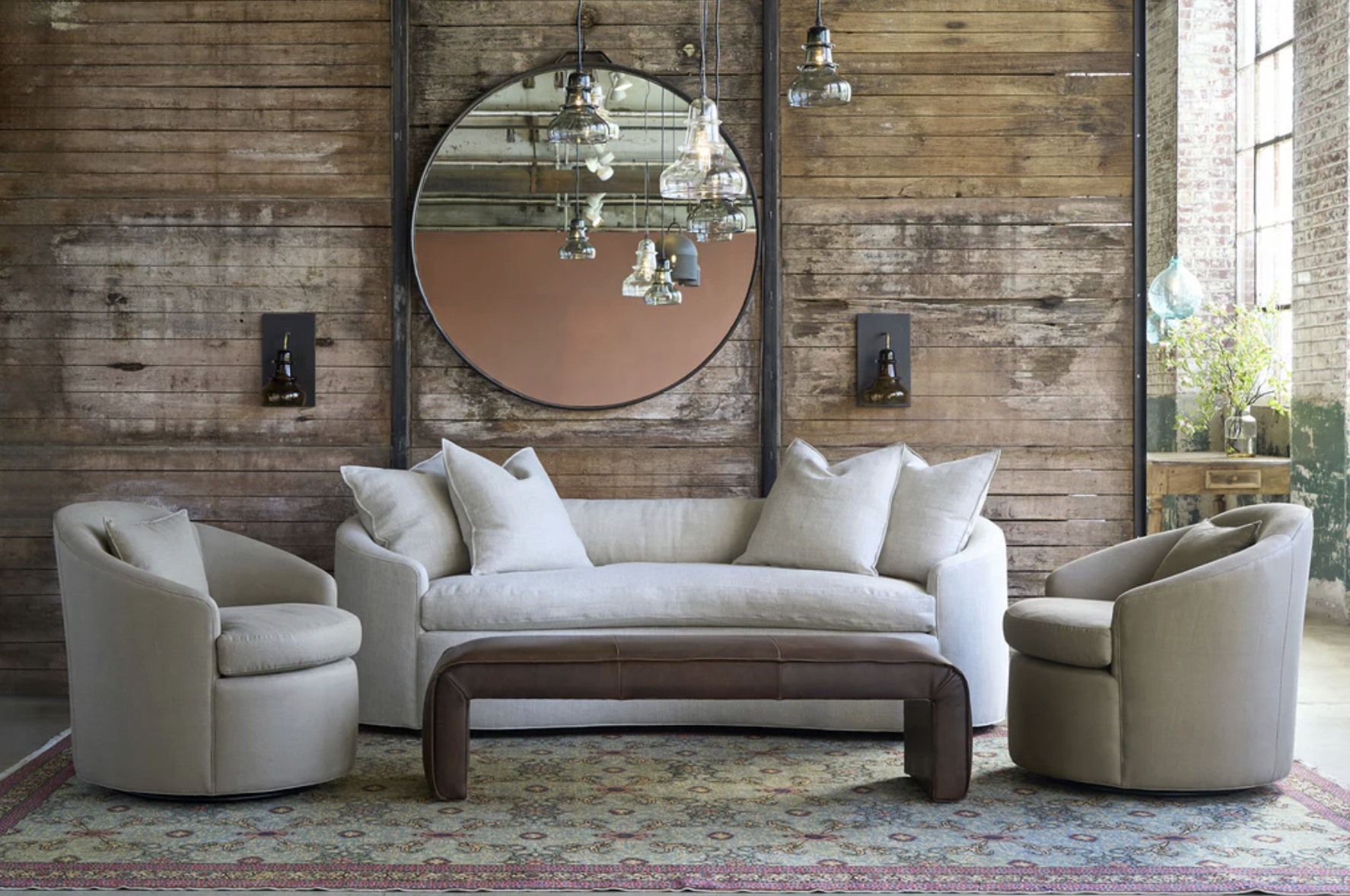 A cozy living room with a white sofa and matching armchairs set against a rustic wooden wall, with a large round mirror, hanging glass pendant lights, and a patterned area rug.
