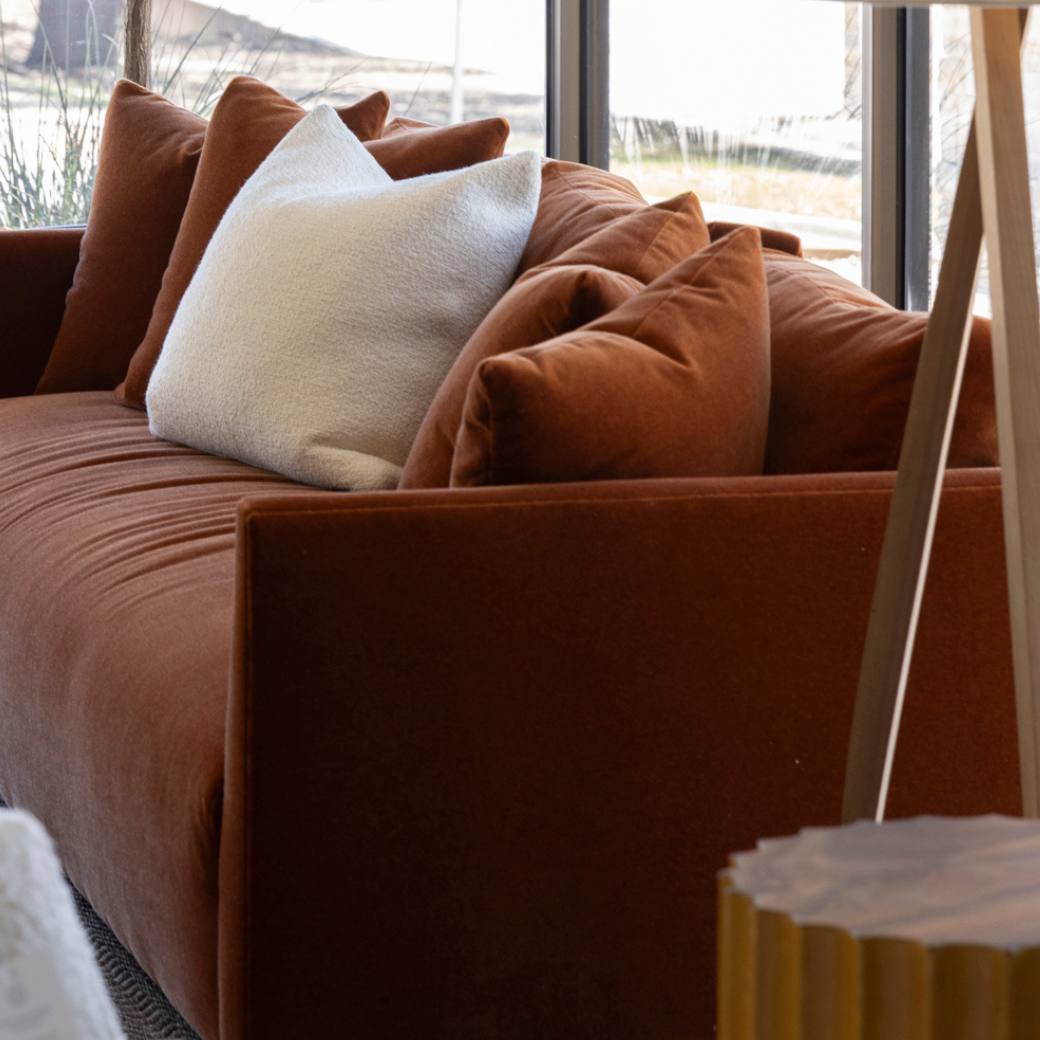 A cozy living room corner with a rust-colored velvet sofa, white and rust-colored pillows, and a large window showing outdoor scenery.
