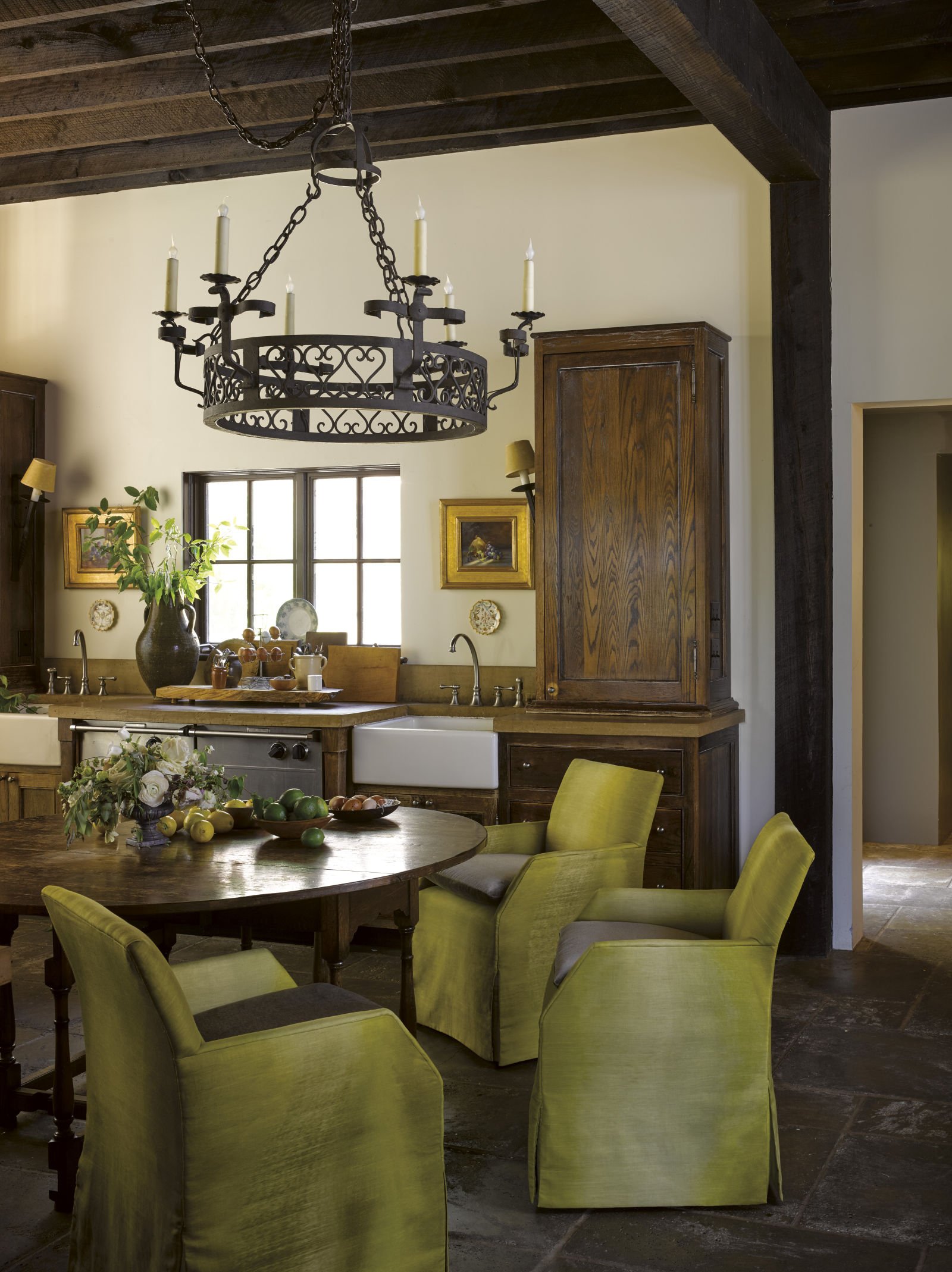 A rustic kitchen with a large wooden dining table surrounded by lime green upholstered chairs, a chandelier hanging from the dark wooden ceiling, and a window with natural light. Decorated with plants, framed artwork, and a fruit bowl.