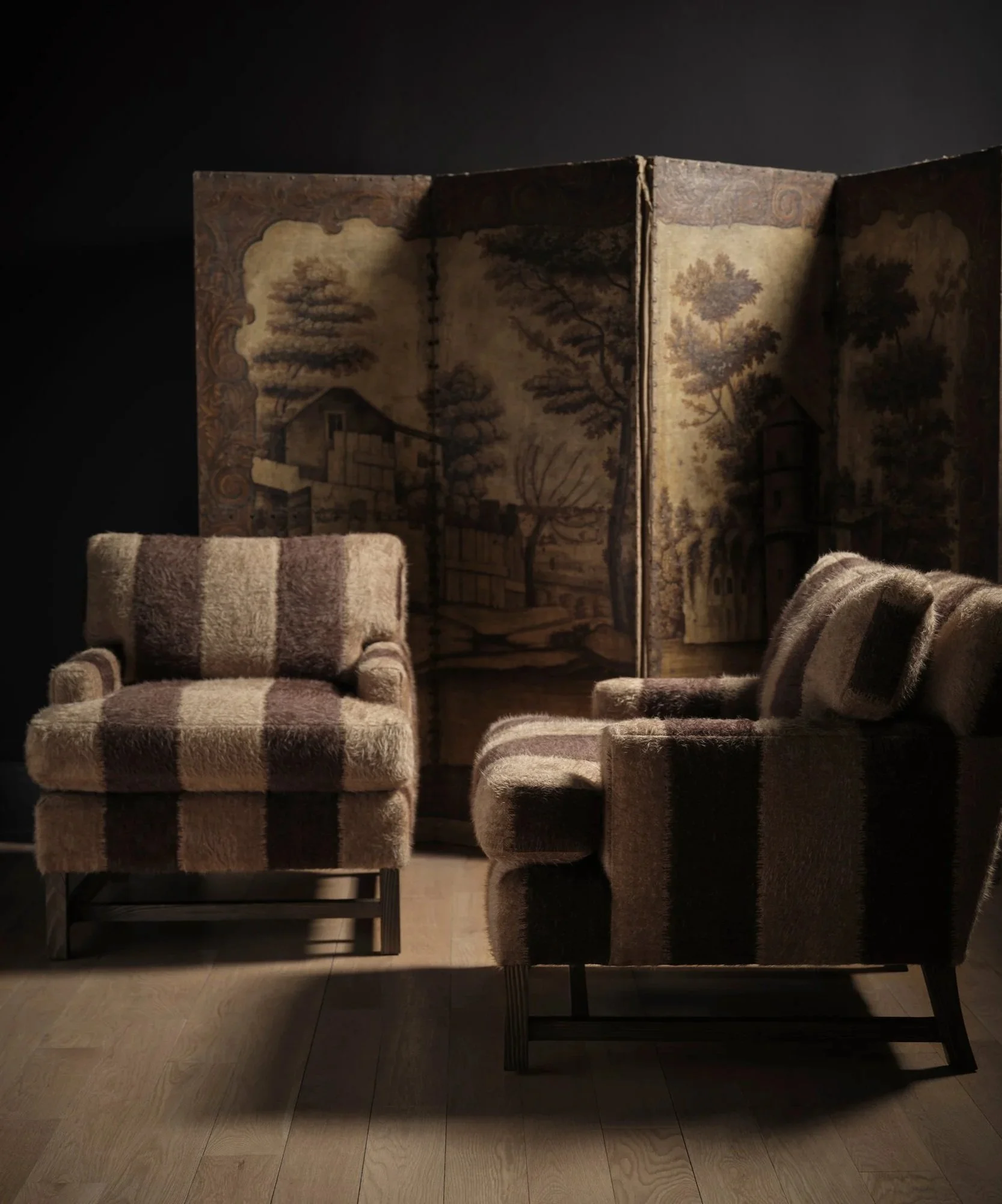A vintage folding screen with a landscape painting of trees and a house, placed behind a cream-colored upholstered armchair with a checkered pattern, and a matching footstool, on wooden flooring against a dark background.