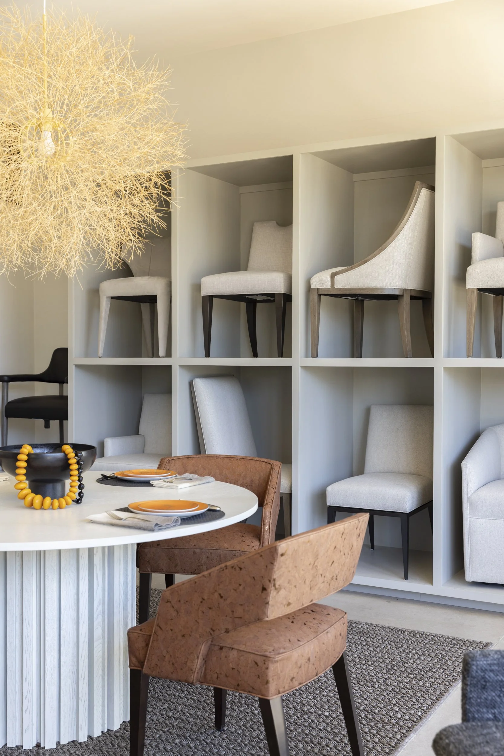 A dining room with a white round table set with orange plates and black and orange beaded jewelry as decor, surrounded by a tan chair and a pink textured chair. Behind the table is a built-in wall shelf displaying various chairs in neutral tones and different styles, with a large modern chandelier hanging from the ceiling.