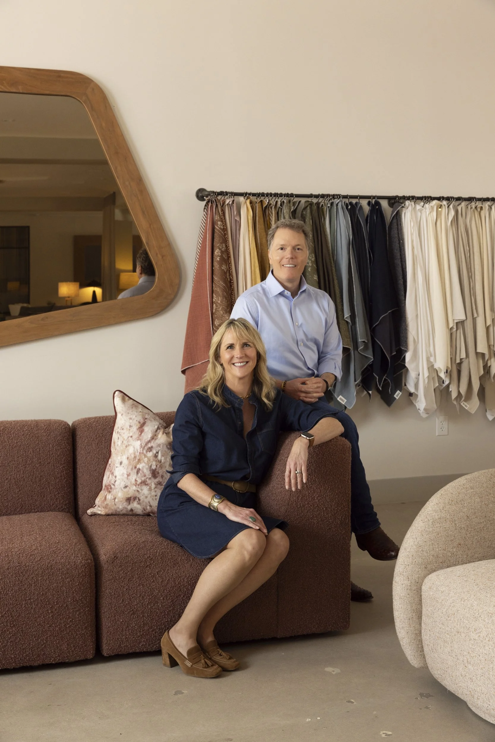A woman sitting on a mauve sofa with a pillow, wearing a dark blue dress and brown heels, has blonde hair and is smiling. Behind her, a man with short brown hair and a light blue shirt stands in front of a fabric display with various colors, and a large wooden-framed mirror is on the wall to the left.
