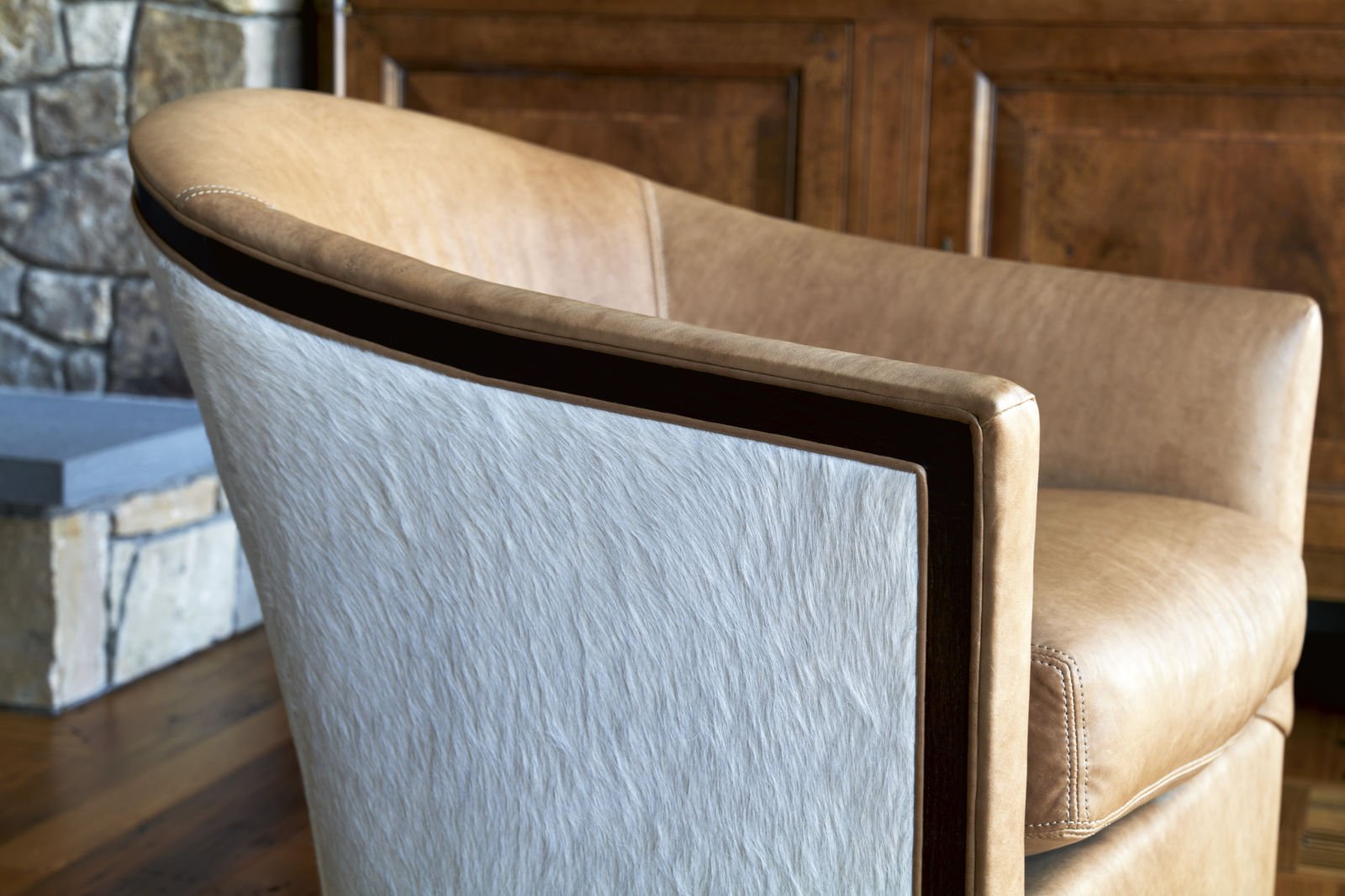 Close-up of an upholstered dining chair with beige fabric, dark wood trim, and a backrest with a curved design in a cozy, rustic room with a stone fireplace and wooden furniture.