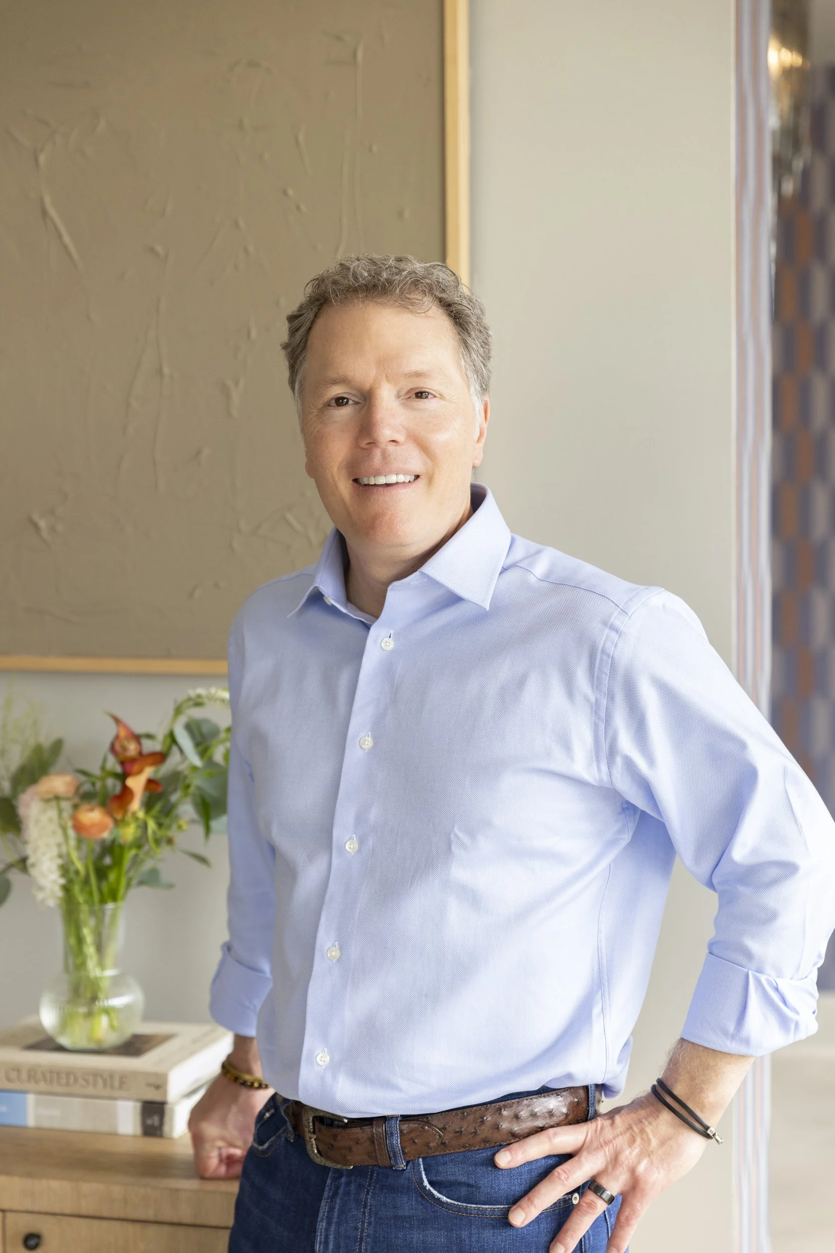 Mid-aged man in blue shirt and jeans standing indoors near a wooden table with a floral bouquet and a stack of books.