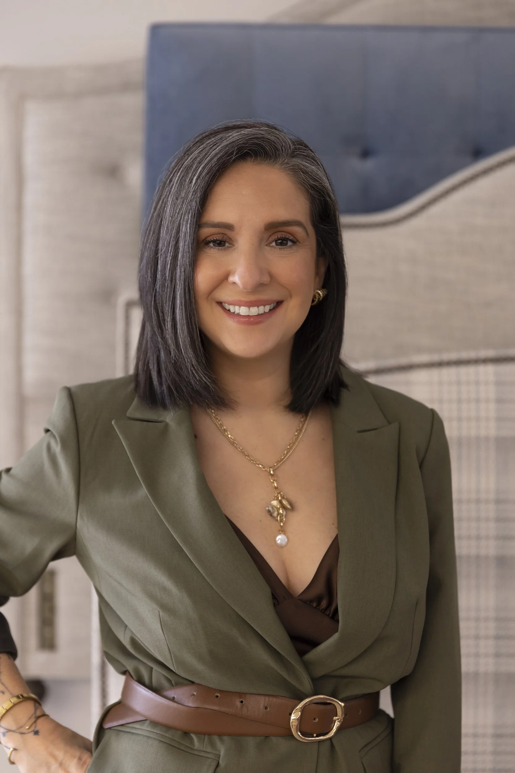 A woman with shoulder-length dark gray hair smiling, wearing a green blazer over a brown blouse, accessorized with gold jewelry including earrings, multiple necklaces, and a brown belt, standing in front of a bed with beige and blue headboards.